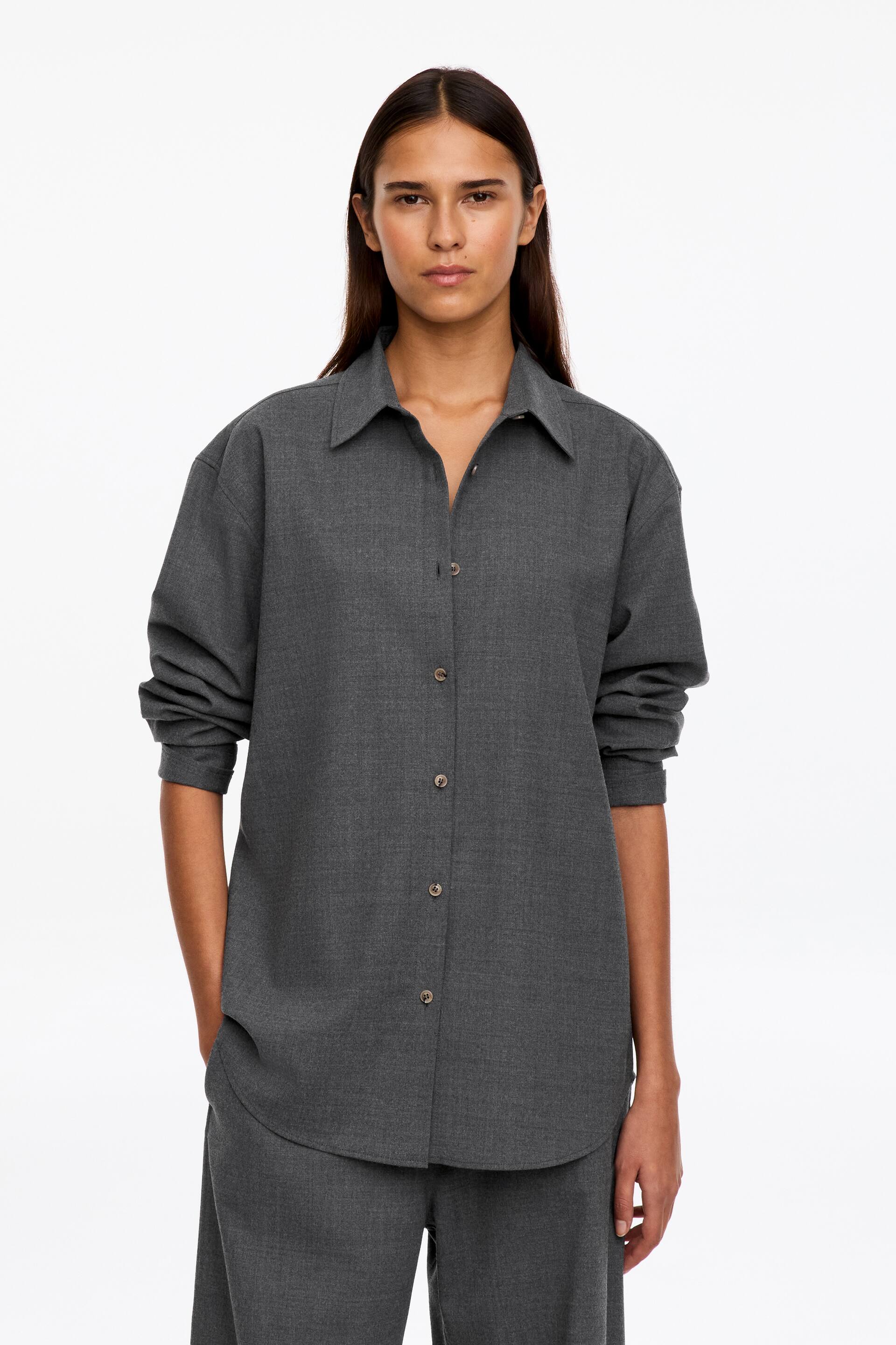 Visualizza l'immagine ingrandita: Una camicia grigia in flanella con abbottonatura frontale, silhouette rilassata, colletto classico e fondo arrotondato è indossata dalla modella. La camicia è abbinata con le maniche arrotolate ed è coordinata con pantaloni grigi assortiti.