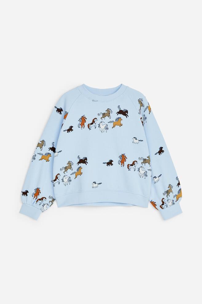 En ljusblÃ¥ sweatshirt med boxig, lÃ¤tt oversized passform visas framifrÃ¥n och framhÃ¤ver ett heltryck med smÃ¥, flerfÃ¤rgade galopperande hÃ¤star. Sweatshirten har lÃ¥nga raglansÃ¤rmar, rund halsringning, ribbade muddar och ribbad fÃ¥ll. - 1