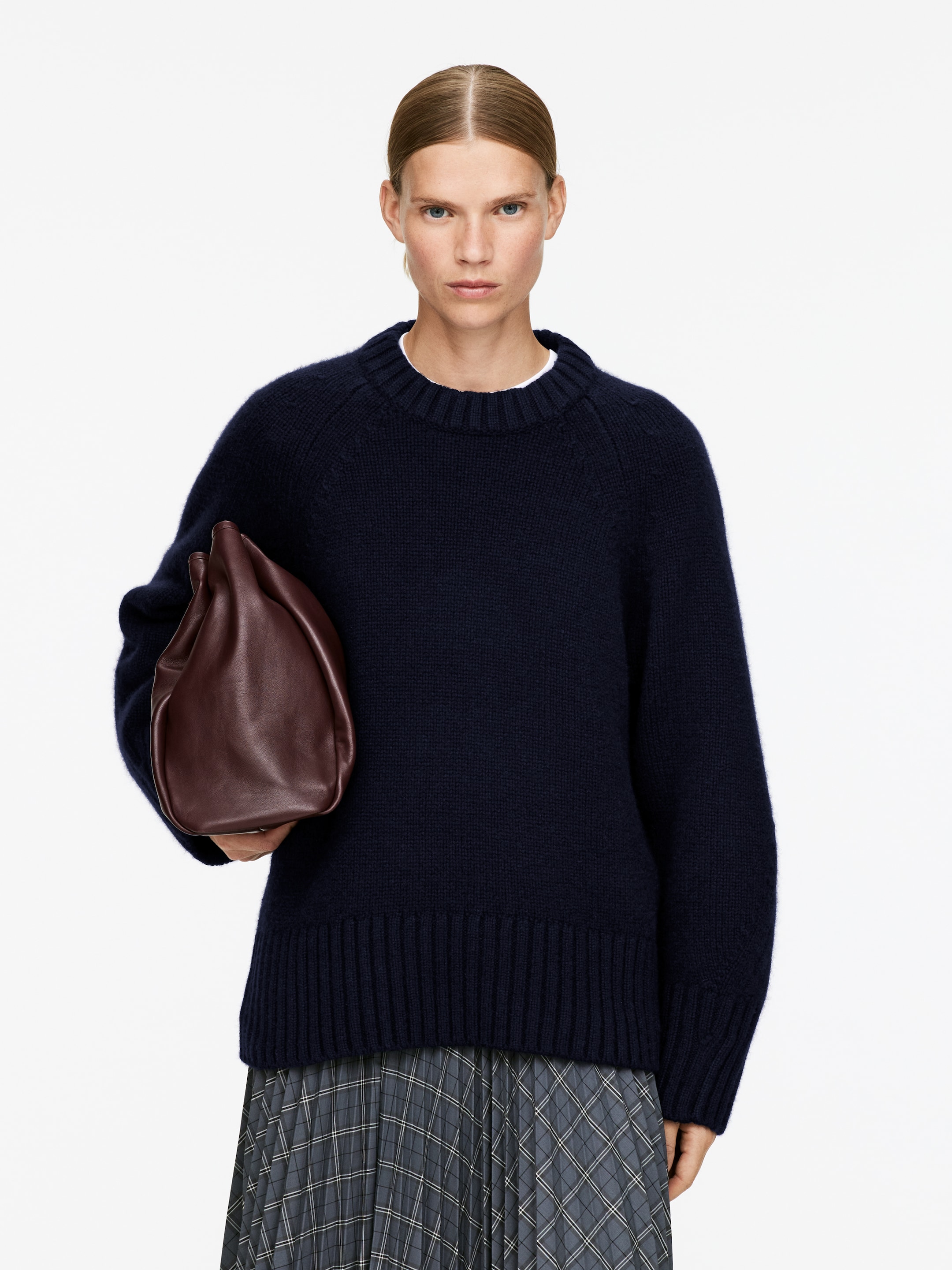 Visualizza immagine più grande: Un maglione girocollo blu navy è mostrato dal davanti, con scollo a coste, polsini a coste e orlo a coste, con maniche raglan visibili. È stratificato sopra una t-shirt bianca, il cui colletto è visibile. Una borsa in pelle morbida marrone scuro con un top raccolto è tenuta nella mano sinistra, leggermente inclinata in avanti.