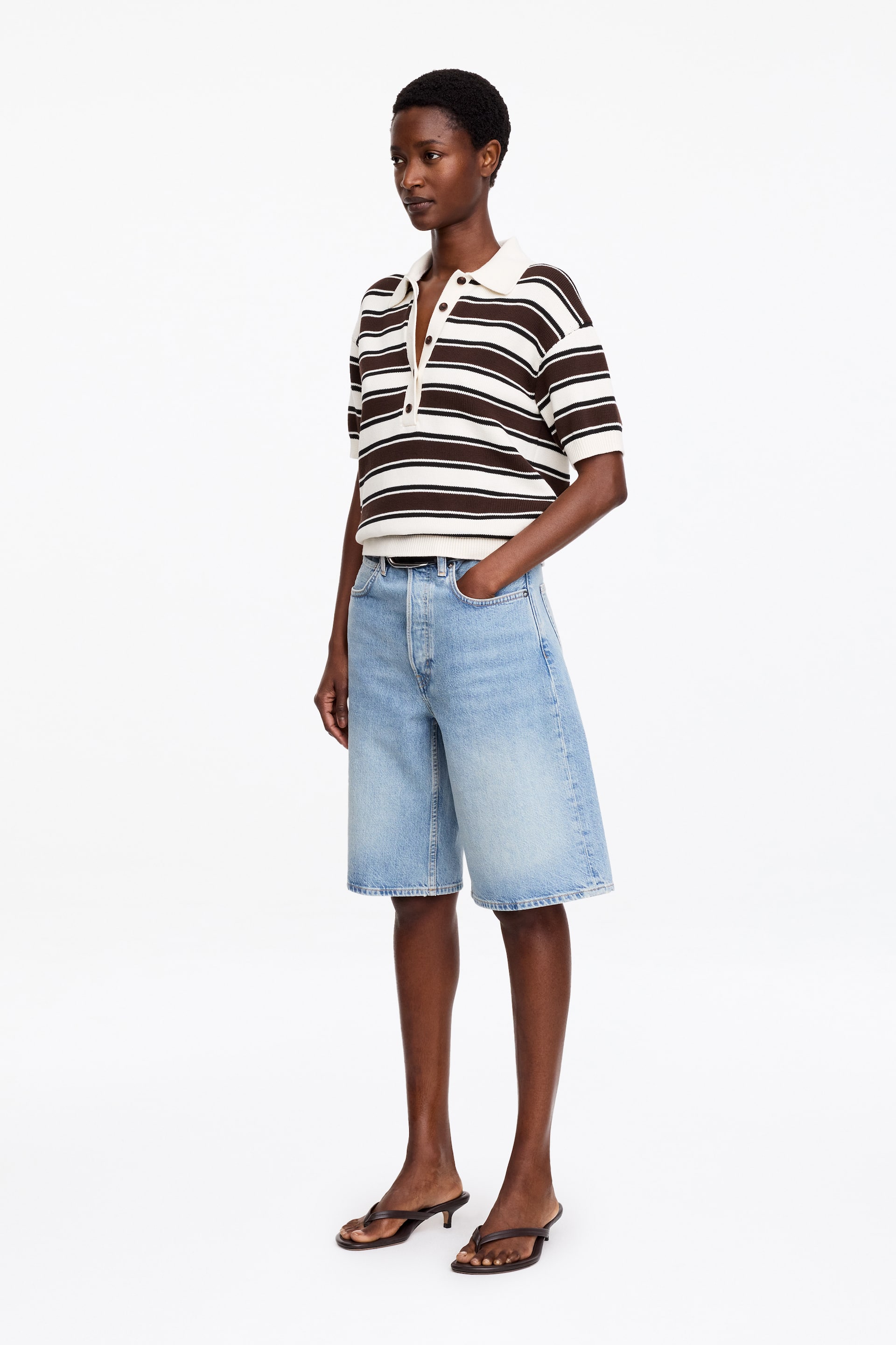 Größeres Bild ansehen: Ein Strick-Poloshirt wird aus einem leichten Seitenwinkel präsentiert, mit breiten braunen und cremeweißen horizontalen Streifen, einem cremeweißen Kragen, kurzen Ärmeln mit gerippten Bündchen und einem gerippten Saum. Dazu werden hell gewaschene Denim-Bermudashorts getragen, die eine entspannte Passform zeigen, mit sichtbarem Knopfverschluss und einem rohen Saum. Ein dünner schwarzer Gürtel betont die Taille, während eine Hand in einer vorderen Tasche ruht.
