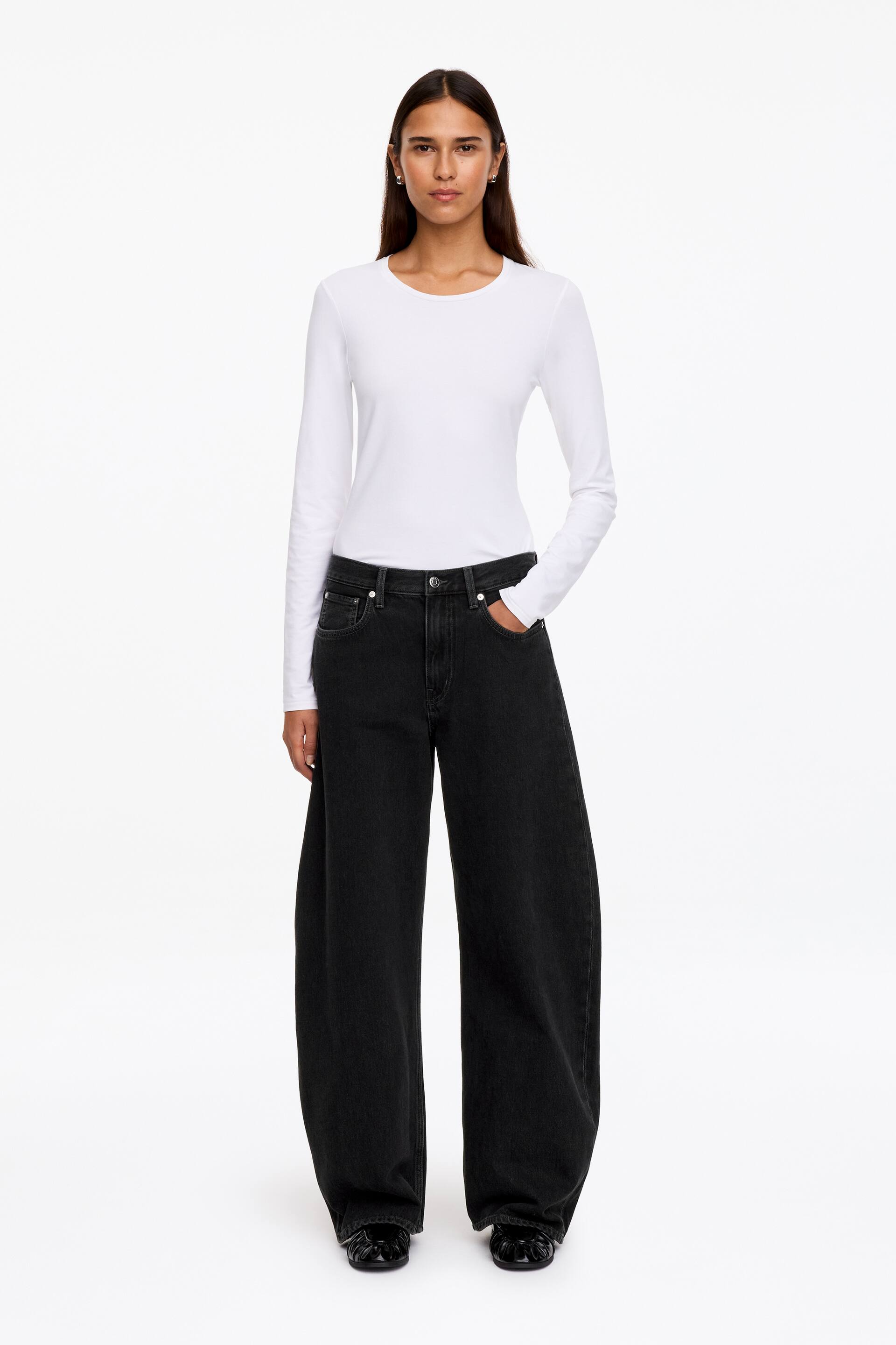 Ver imagem maior: Jeans pretos de perna afunilada, com silhueta descontraída e curvada e cintura média, estão a ser usados pela modelo. A modelo está de pé com uma mão metida no bolso da frente, combinando as jeans com um top branco de manga comprida metido por dentro.