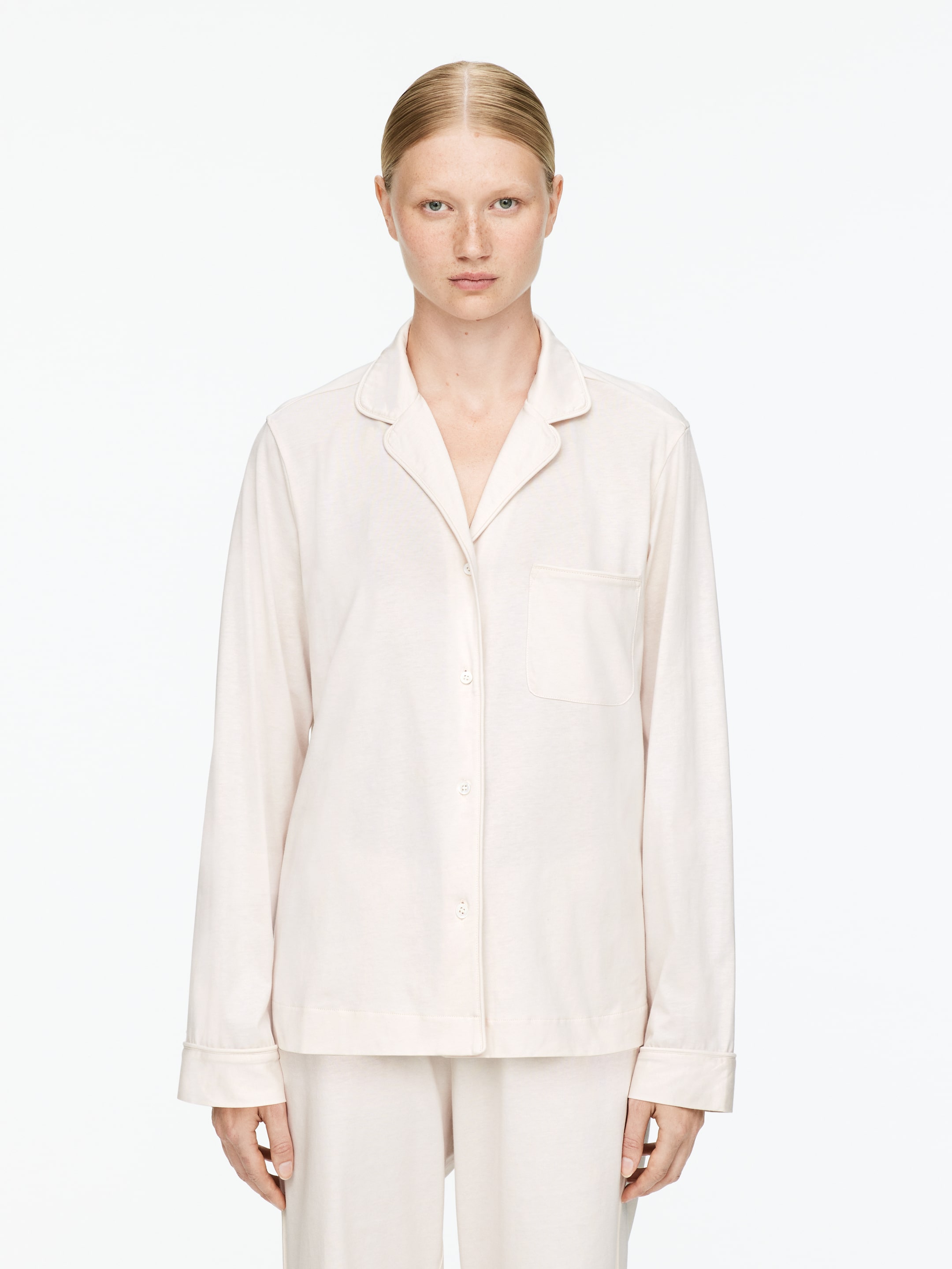 Agrandir l'image: Un ensemble de pyjama crème clair est présenté en vue de face, comprenant un haut à manches longues avec des revers crantés, une fermeture à boutons sur le devant, une poche poitrine unique et des détails de passepoil subtils, assorti à un pantalon ample assorti.