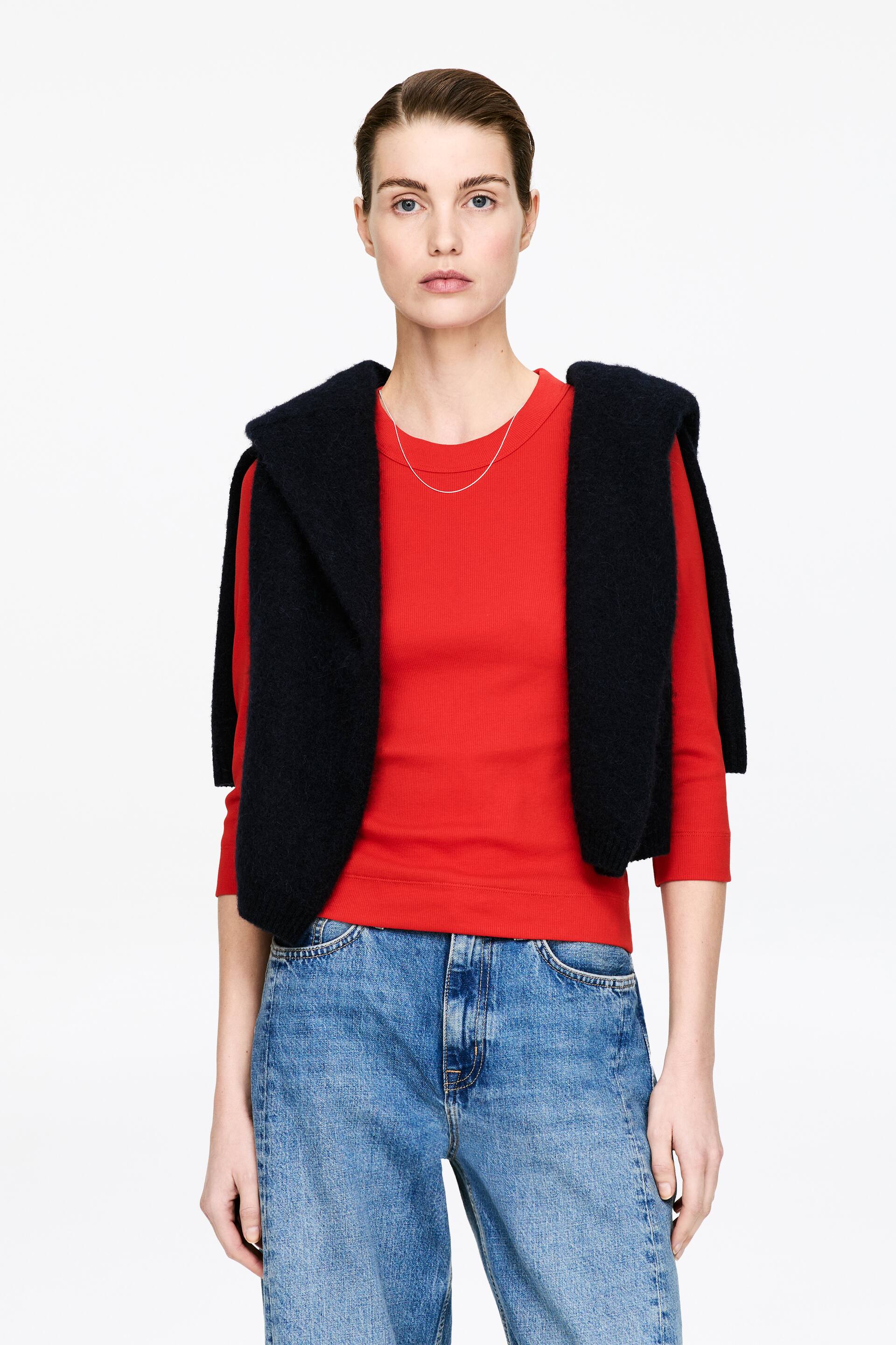Visualizza immagine più grande: Un top rosso a coste con maniche a tre quarti e scollo rotondo. Un maglione scuro e testurizzato è drappeggiato sulle spalle, mentre una sottile collana d'argento riposa sullo scollo.