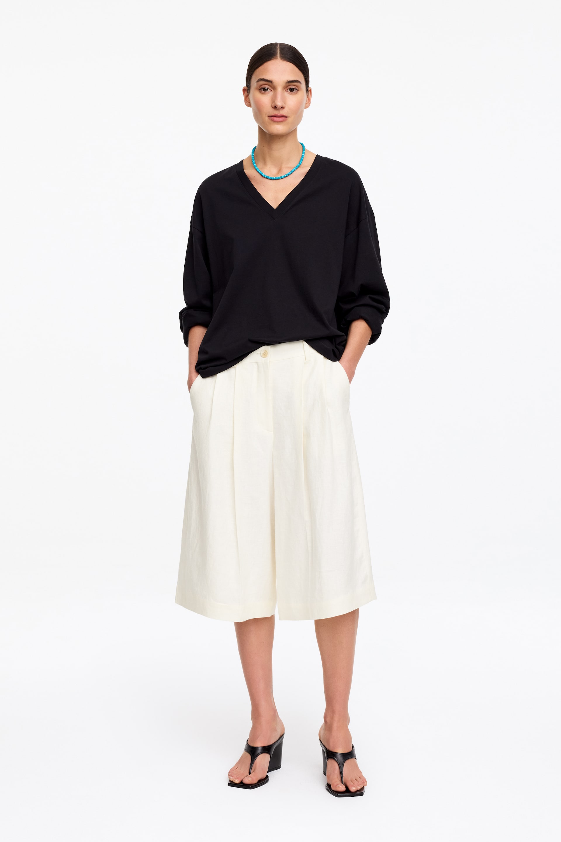 Grotere afbeelding bekijken: Off-white linnen shorts met voorplooien en geperste vouwen. Het model draagt de shorts, met de handen in de schuin geplaatste zijzakken, en een enkele witte knoop is zichtbaar in de taille.