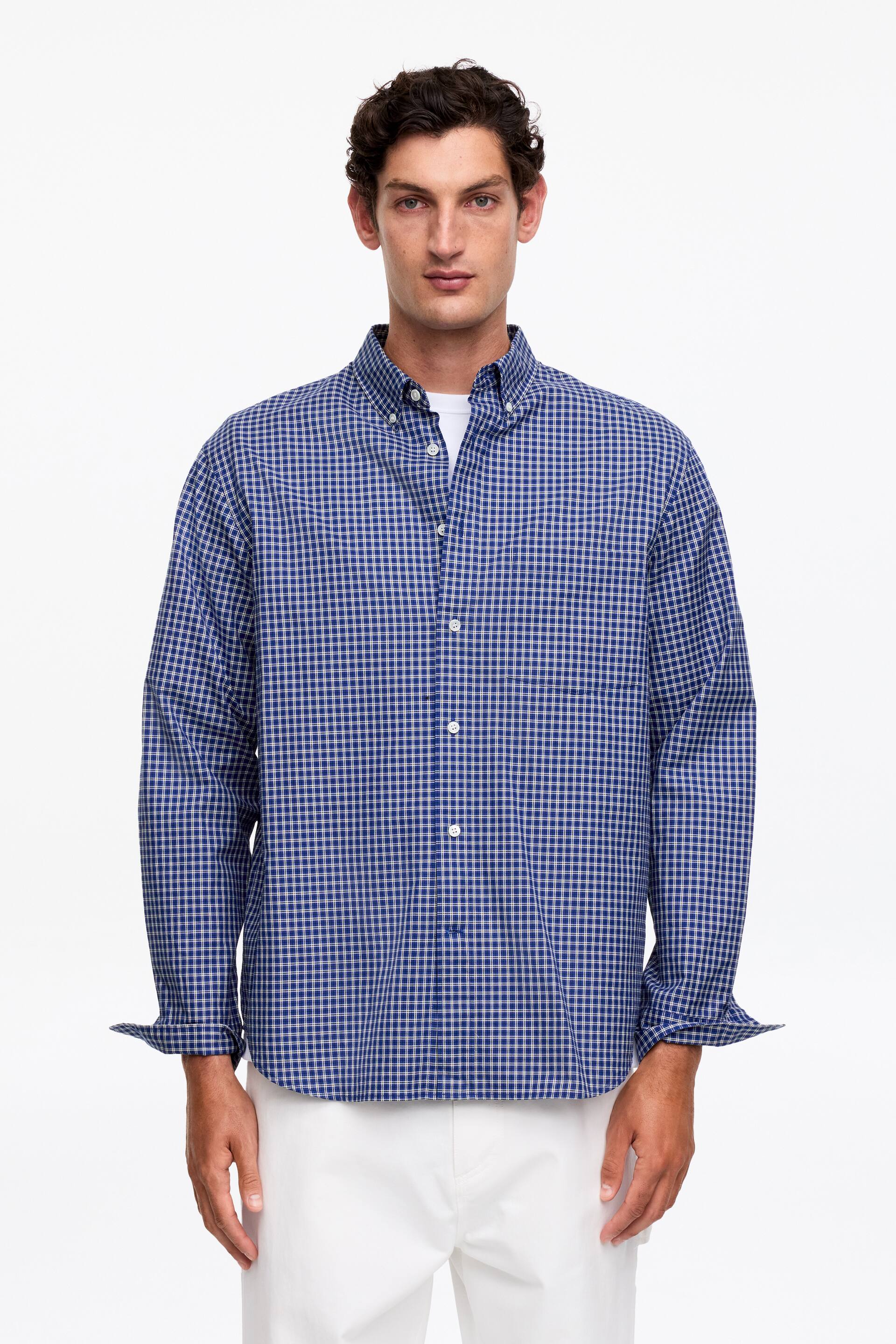 Visa större bild: En skjorta med ett blått och vitt smårutigt mönster bärs framifrån, med en button-down krage och vita knappar längs den främre knappslån. Dess långa ärmar är uppvikta vid manschetterna, och en vit t-shirt syns under den öppna kragen.