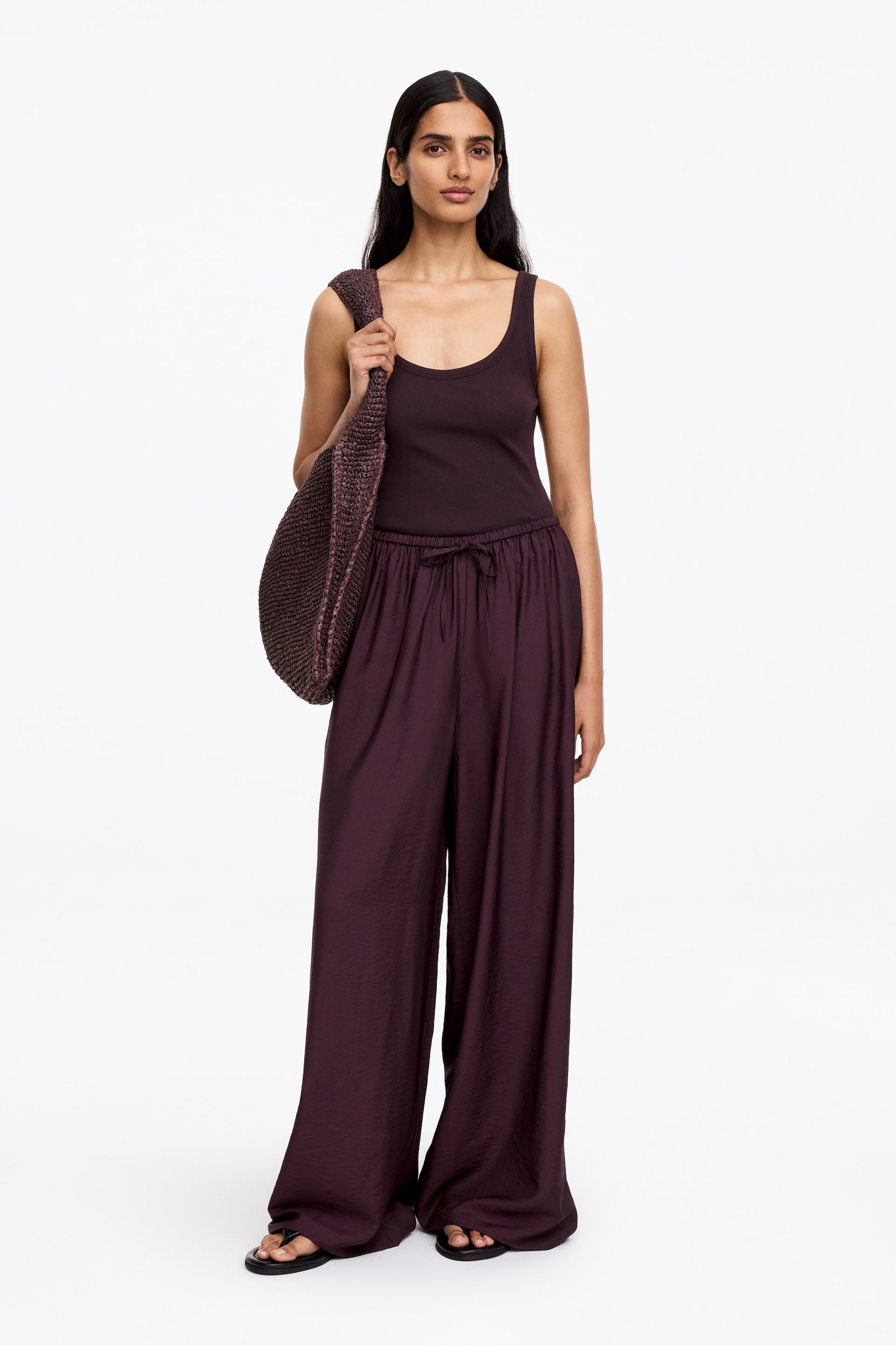Agrandir l'image: Pantalon violet foncé, confectionné dans un tissu lisse et fluide, présentant une coupe ample et une silhouette à jambes larges avec une taille froncée et un cordon de serrage visible. Le mannequin porte le pantalon, associé à un débardeur assorti et des tongs noires, et porte un sac tissé sur une épaule.