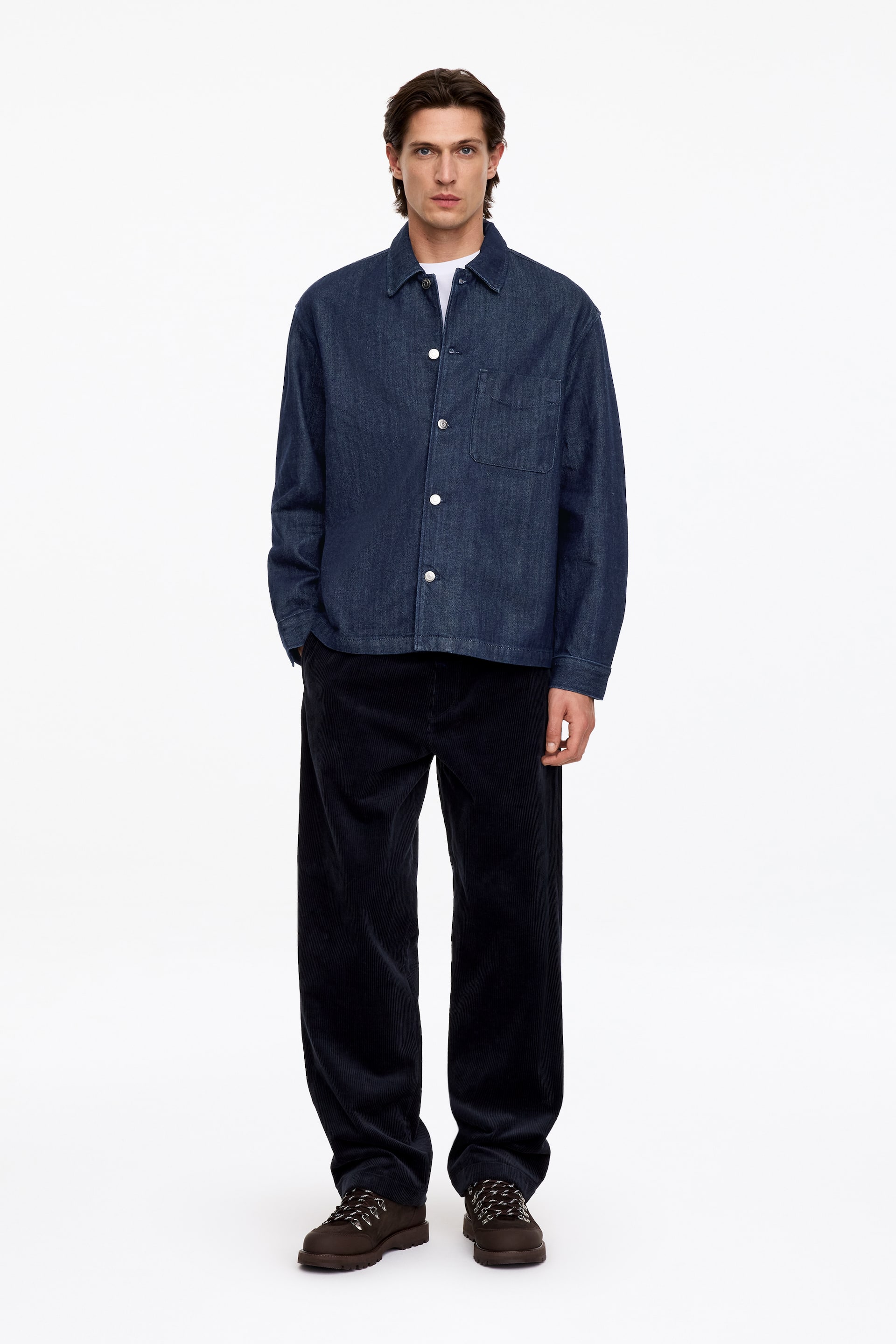 Agrandir l'image: Une surchemise en denim bleu foncé avec un col classique, une fermeture boutonnée sur le devant et une seule poche plaquée sur la poitrine est portée par le mannequin. La surchemise est superposée à un t-shirt blanc et associée à un pantalon en velours côtelé foncé à jambes larges.