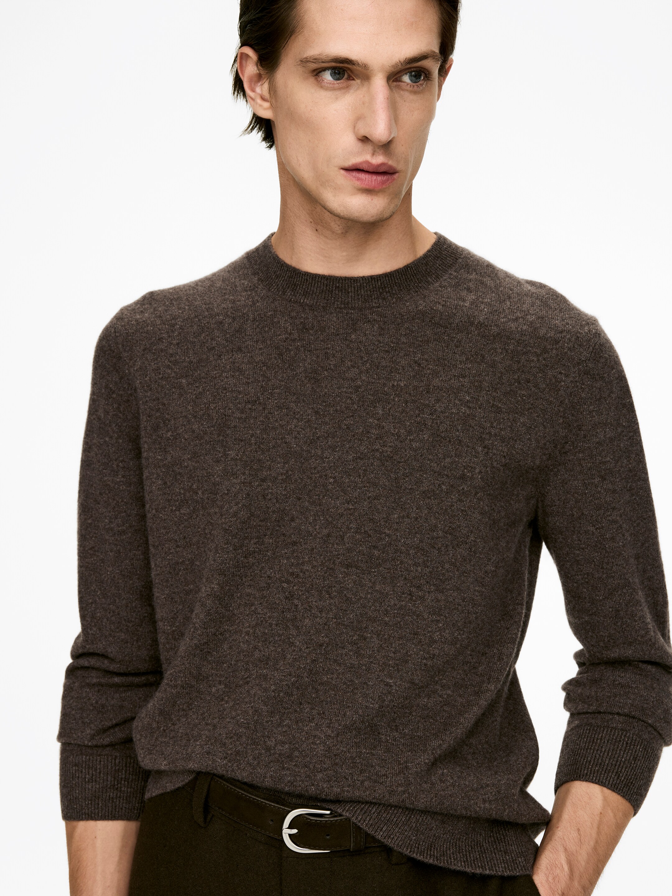 View larger image: A dark brown crew-neck sweater is being presented, angled slightly to the right. It is featuring a fine knit texture, ribbed cuffs, and a ribbed hem. The left side of the sweater is being subtly tucked into dark trousers, revealing a silver-buckled belt.