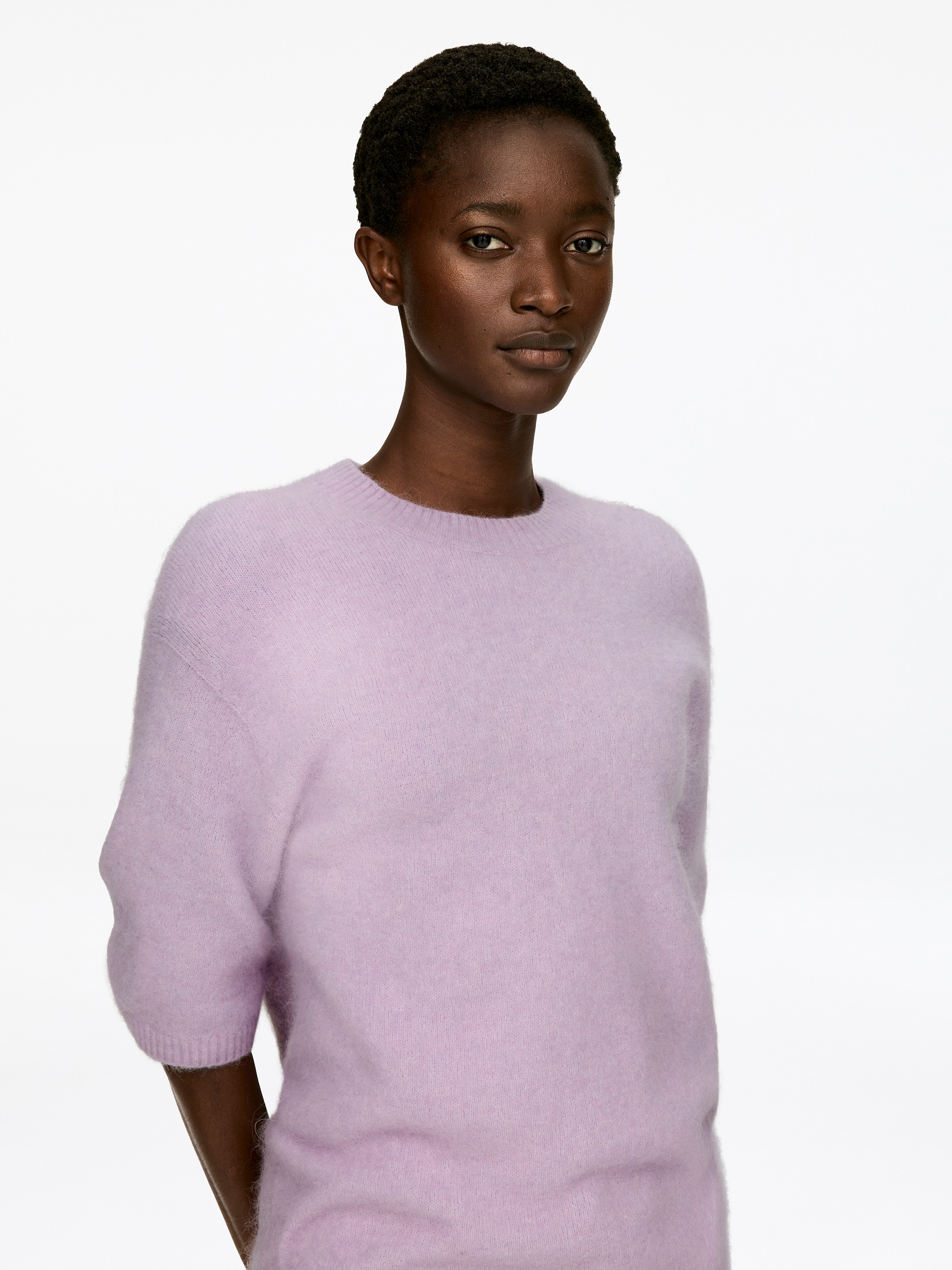 Visualizza immagine più grande: Un maglione a girocollo color lavanda viene indossato, rivolto in avanti, e presenta maniche corte con polsini a coste. Presenta una texture lavorata a maglia morbida e pelosa e una vestibilità rilassata.