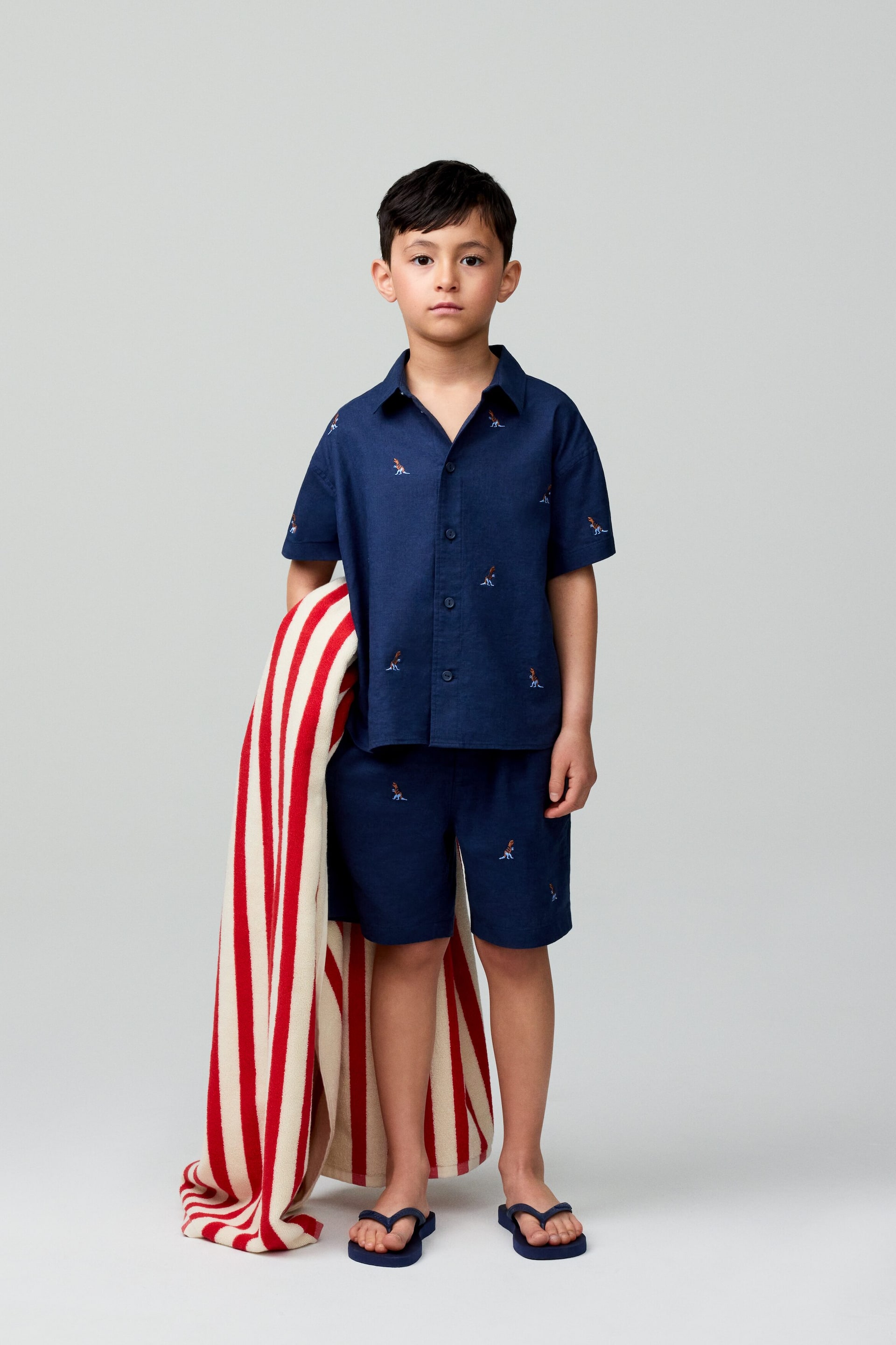 Agrandir l'image: Un ensemble de chemise à manches courtes et short assortis pour enfant en bleu marine est présenté de face. La chemise arbore un col classique et une fermeture à boutons, et à la fois la chemise et le short affichent de petites figures de surfeurs brodées.