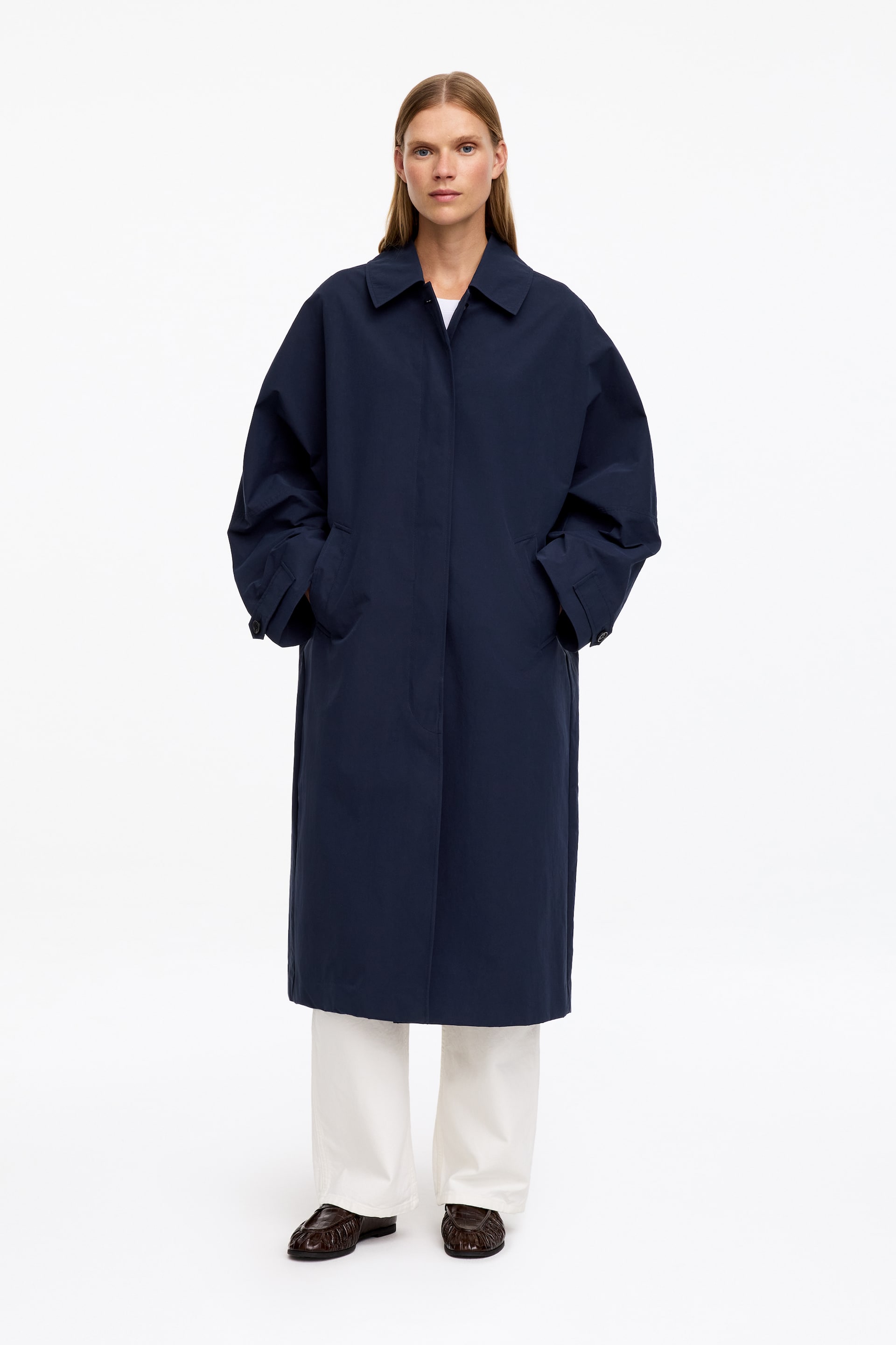 Agrandir l'image: Un car coat bleu marine, doté d’un col classique, d’une silhouette décontractée et d’une longueur mi-mollet, est porté par le mannequin. Les mains du mannequin reposent dans les poches passepoilées latérales du manteau. Le car coat est associé à un haut blanc et à un pantalon large de couleur claire.