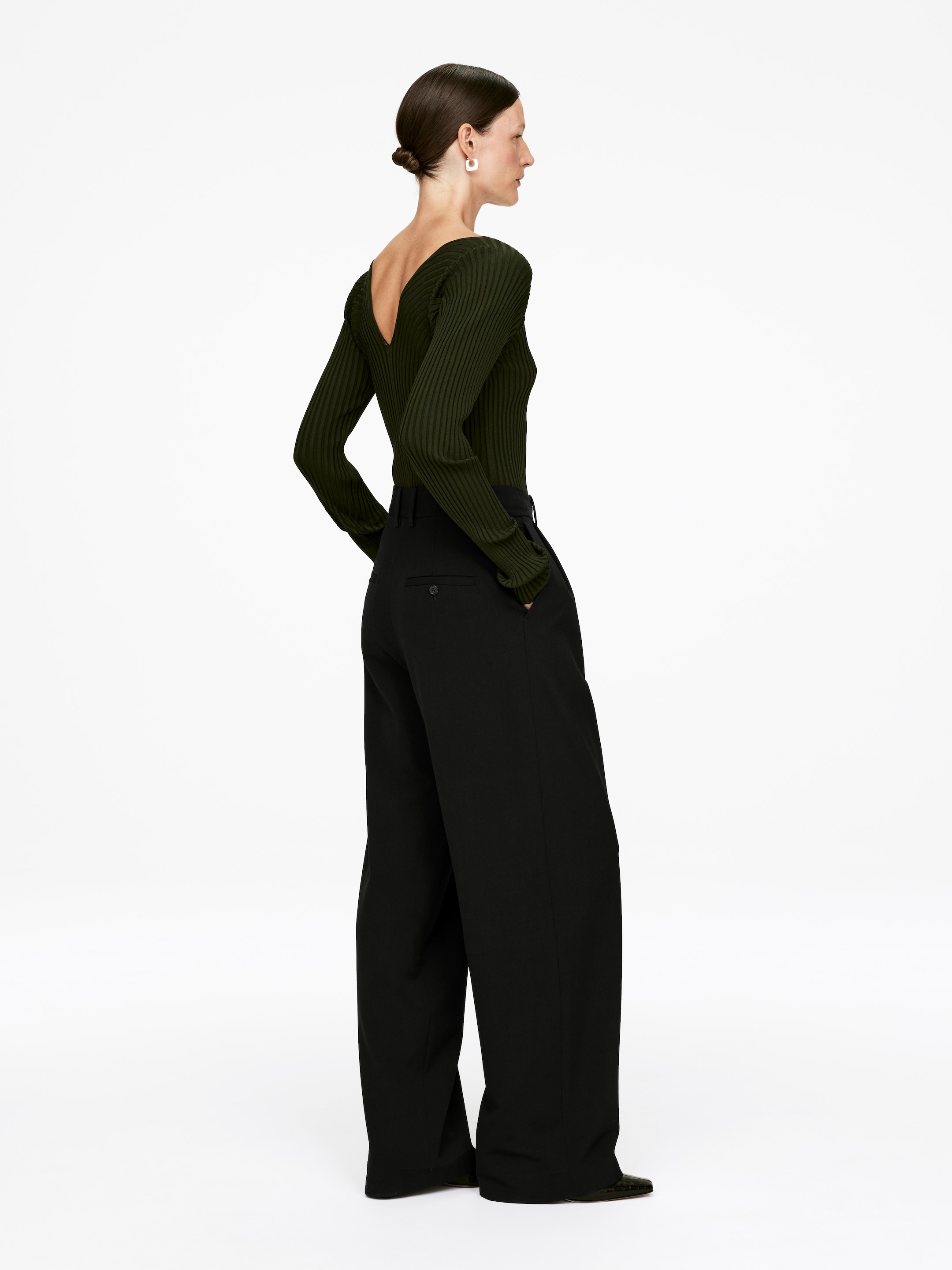 View larger image: A dark green ribbed knit top, featuring a deep V-back neckline and long, fitted sleeves, is being worn with high-waisted black wide-leg trousers. The trousers are displaying a relaxed fit with a visible button closure, and the ensemble is being presented from a rear-side angle.