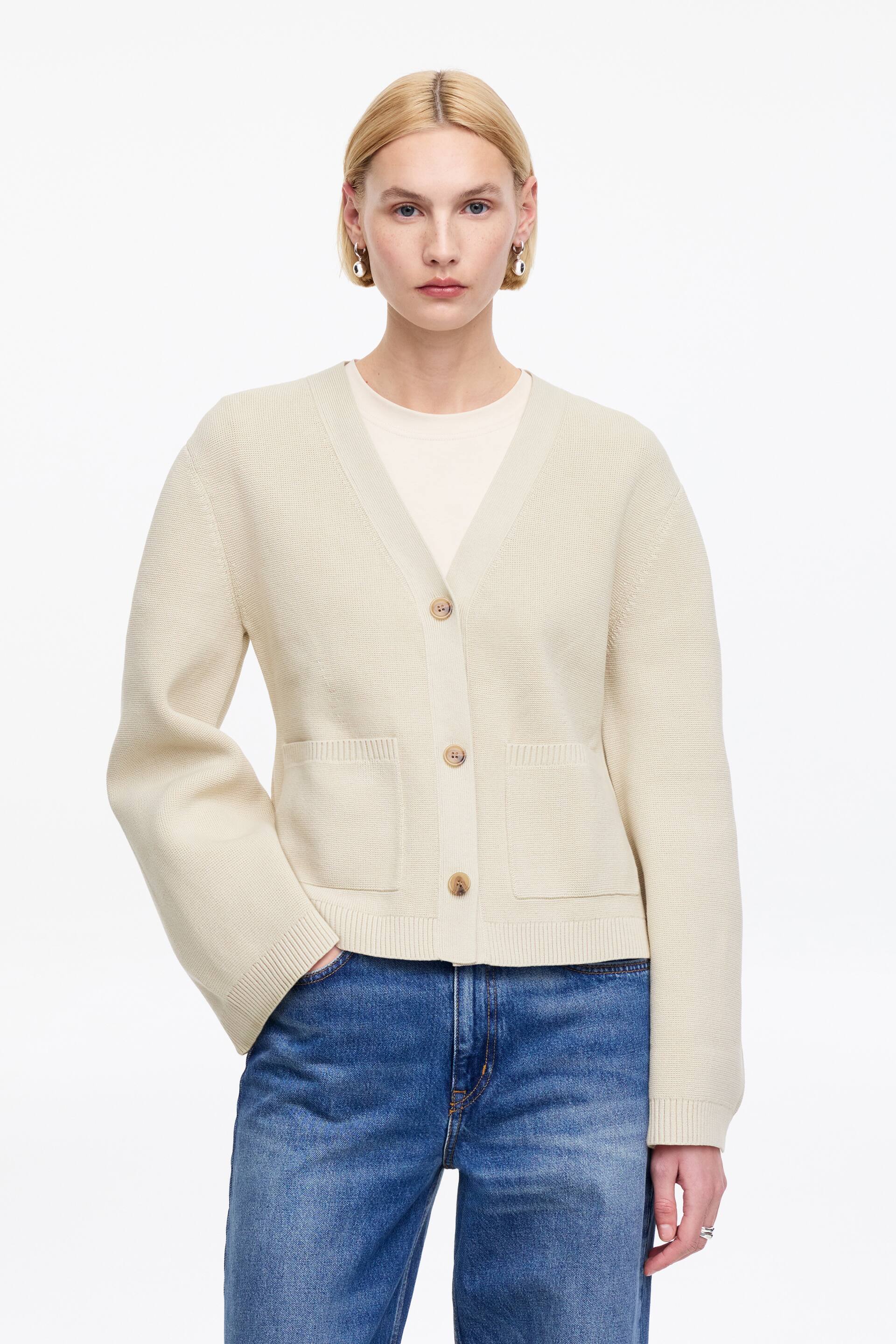 Größeres Bild ansehen: Ein cremefarbener V-Ausschnitt-Cardigan wird über einem hellen T-Shirt getragen und ist nach vorne gerichtet. Er verfügt über drei hellbraune Knöpfe an der Vorderseite, zwei rechteckige aufgesetzte Taschen in der Nähe des Saums sowie gerippte Bündchen und einen gerippten Saum.