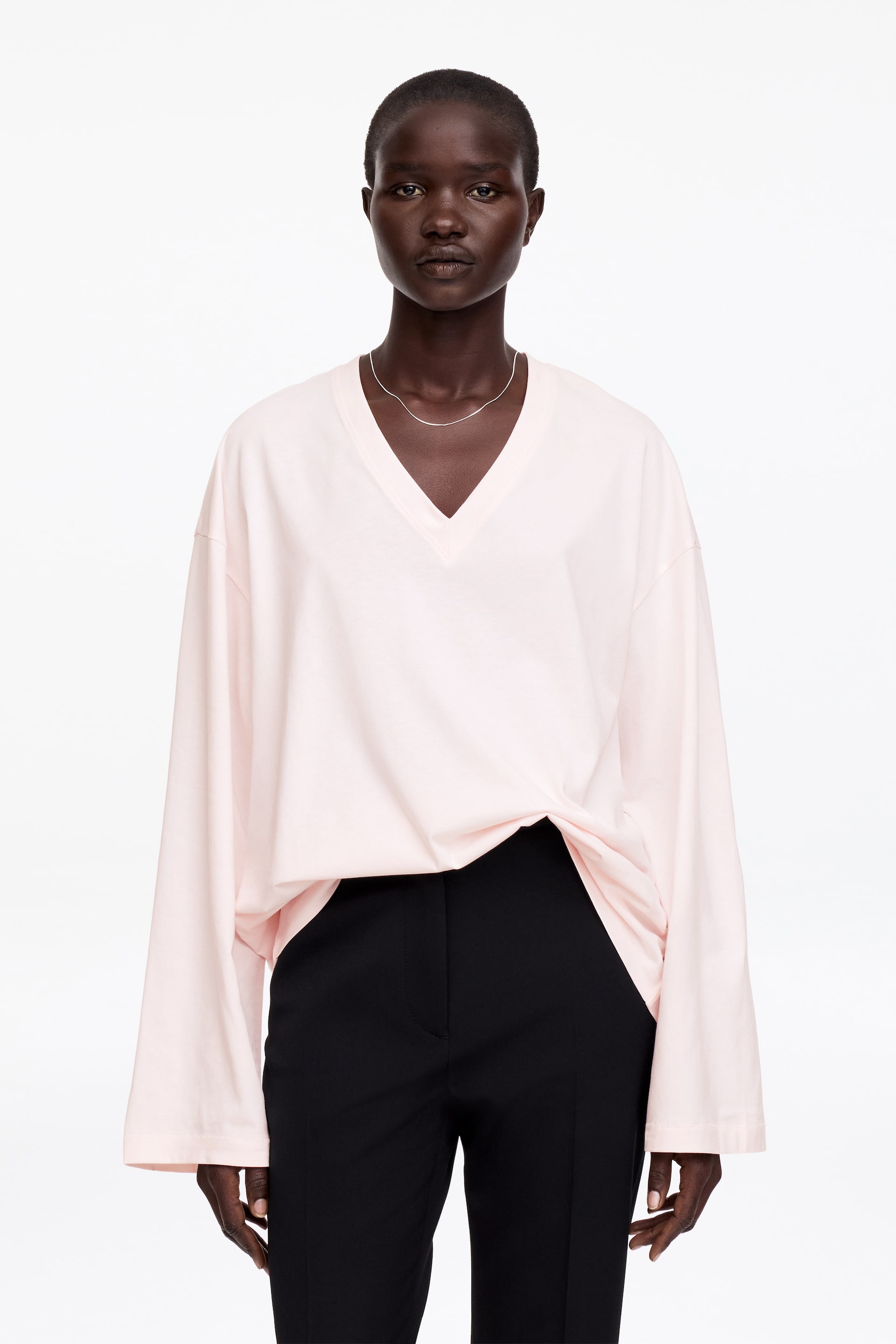 Visa större bild: En ljusrosa V-ringad långärmad topp visas, med en överdimensionerad, avslappnad passform och lösa, draperade ärmar. Toppen är vänd framåt, med ett tunt silverhalsband vilande på bröstet ovanför V-ringningen, och är delvis instoppad i svarta byxor på ena sidan.