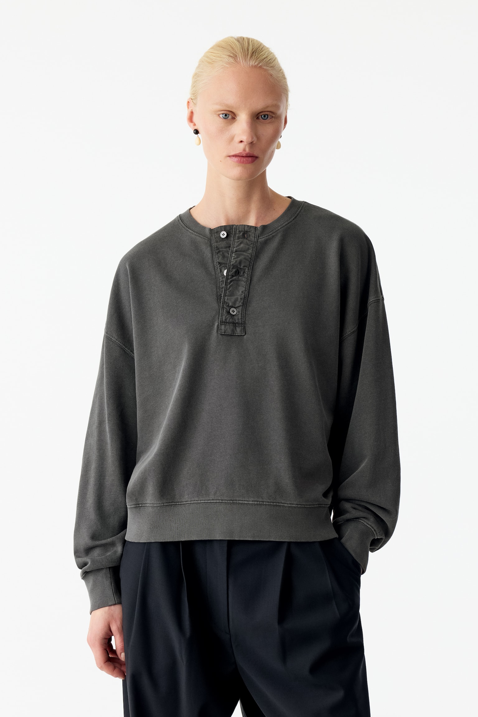 Een verweerde houtskoolgrijze Henley-sweatshirt met een geknoopte knoopsluiting, afhangende schouders en geribde boorden wordt gedragen door het model. De sweatshirt met relaxte pasvorm is gestyled met donker geplooide broek. - 1