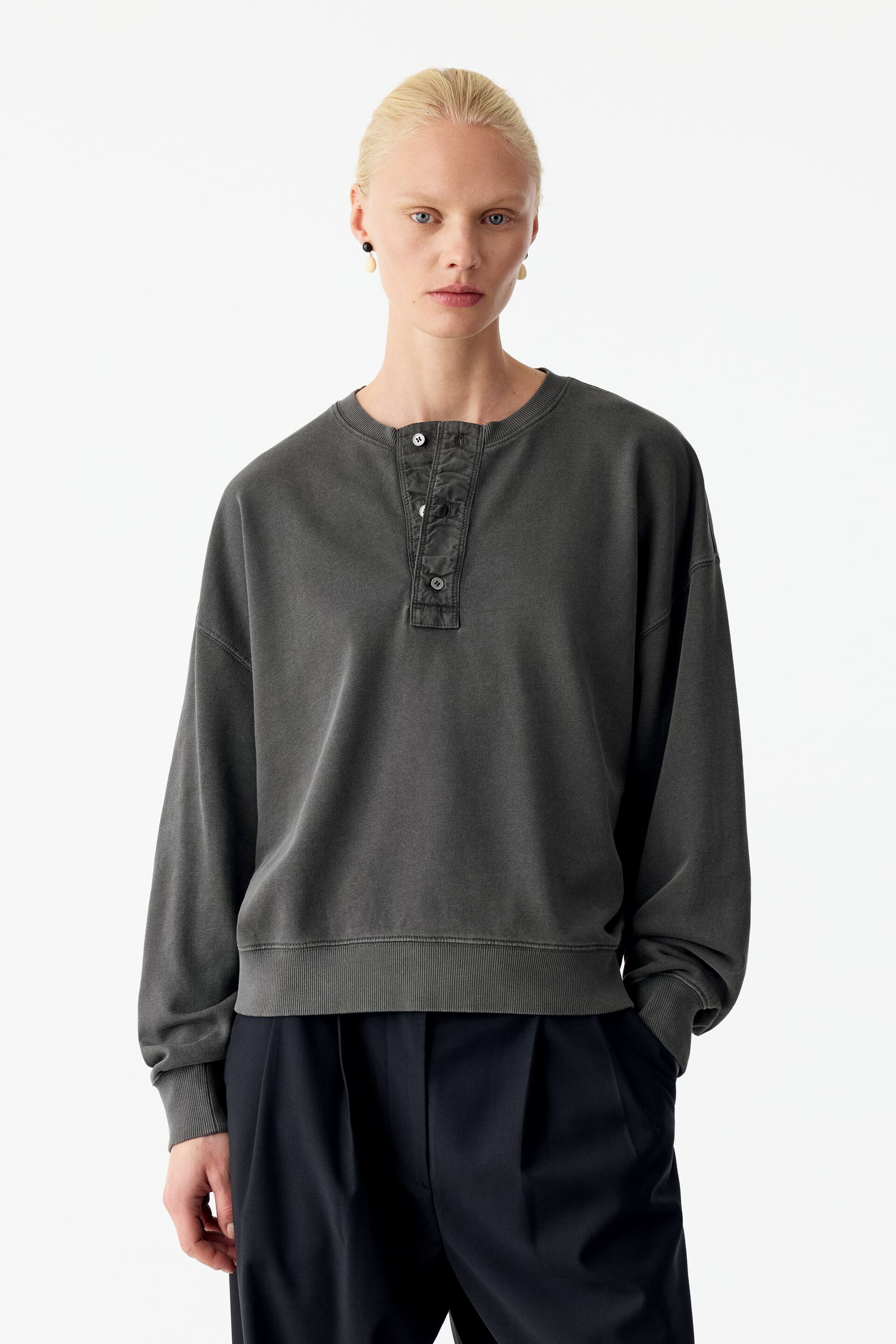 Ver imagen más grande: La modelo lleva una sudadera Henley en gris carbón descolorido con tapeta abotonada, hombros caídos y ribetes de canalé. La sudadera de corte relajado se combina con pantalones oscuros de pinzas.