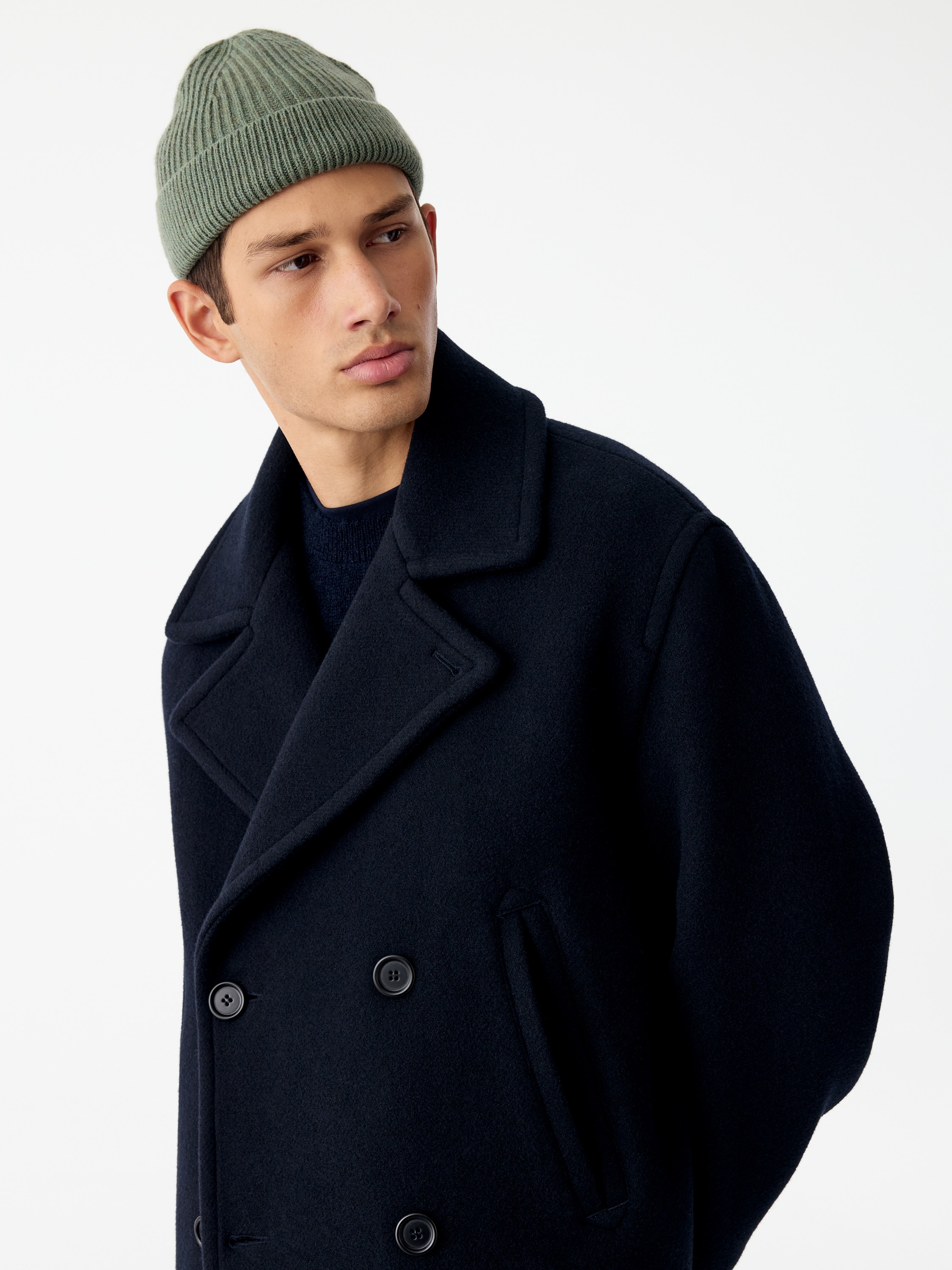 A ribbed knit beanie in a muted sage green is facing the front, displaying a rounded crown and a wide, folded-up brim with a vertical ribbed pattern.