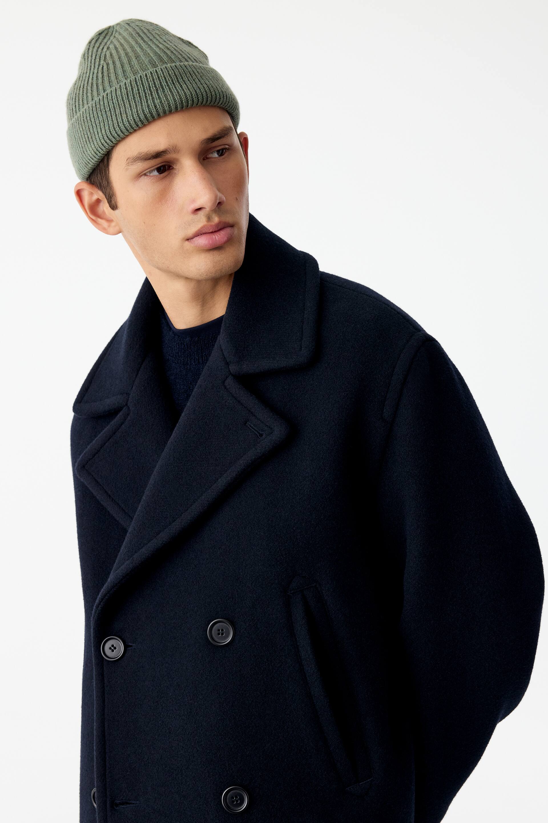 View larger image: A light green ribbed-knit beanie with a folded brim is being worn by the model. The short-style beanie is styled with a dark navy double-breasted wool coat.