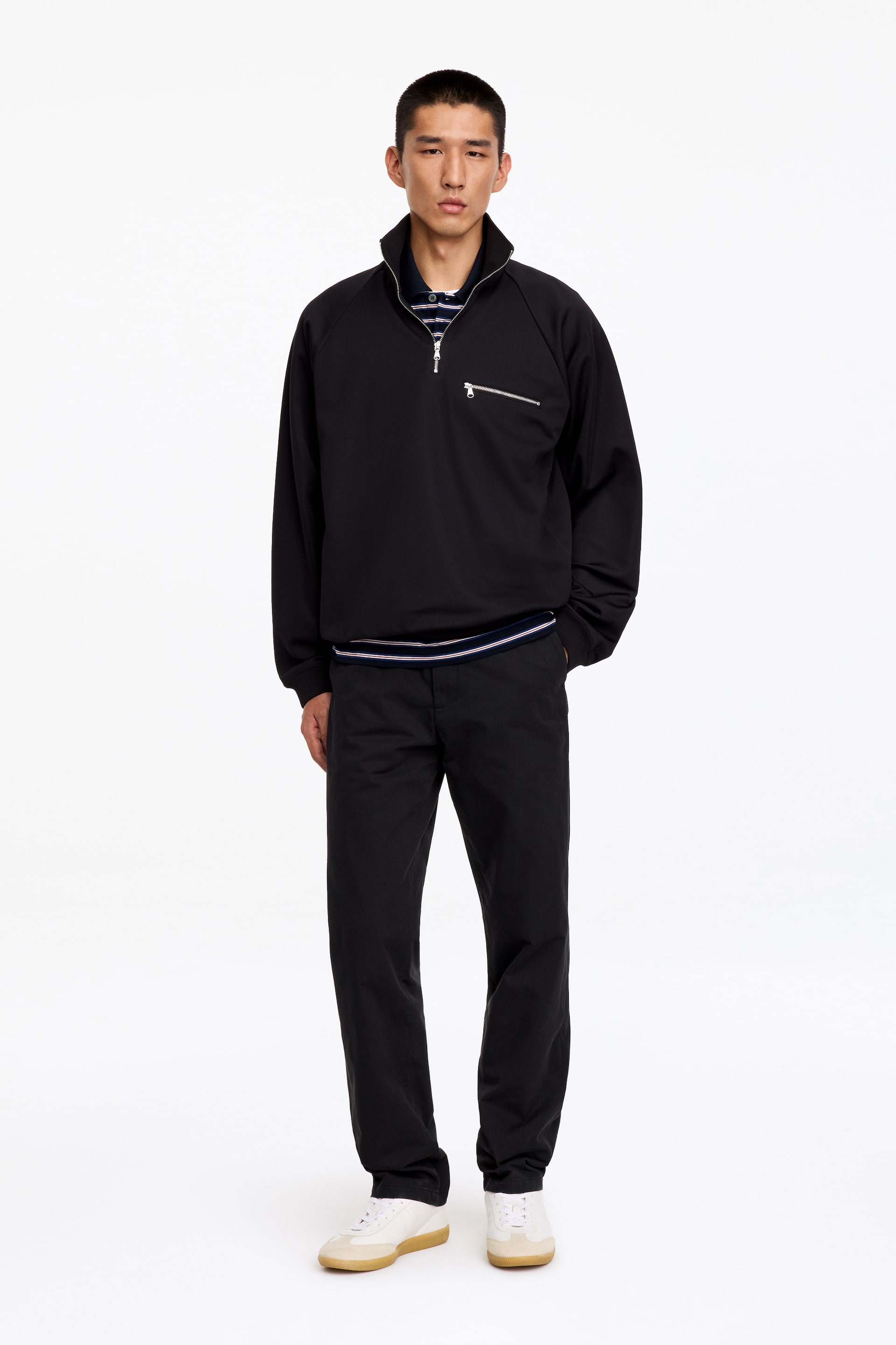 Agrandir l'image: Le mannequin porte un chino noir en coton à coupe droite classique, avec une jambe droite, et se tient debout, une main glissée dans la poche avant. Le pantalon est associé à un sweat-shirt noir à demi-zip et à des baskets blanches.