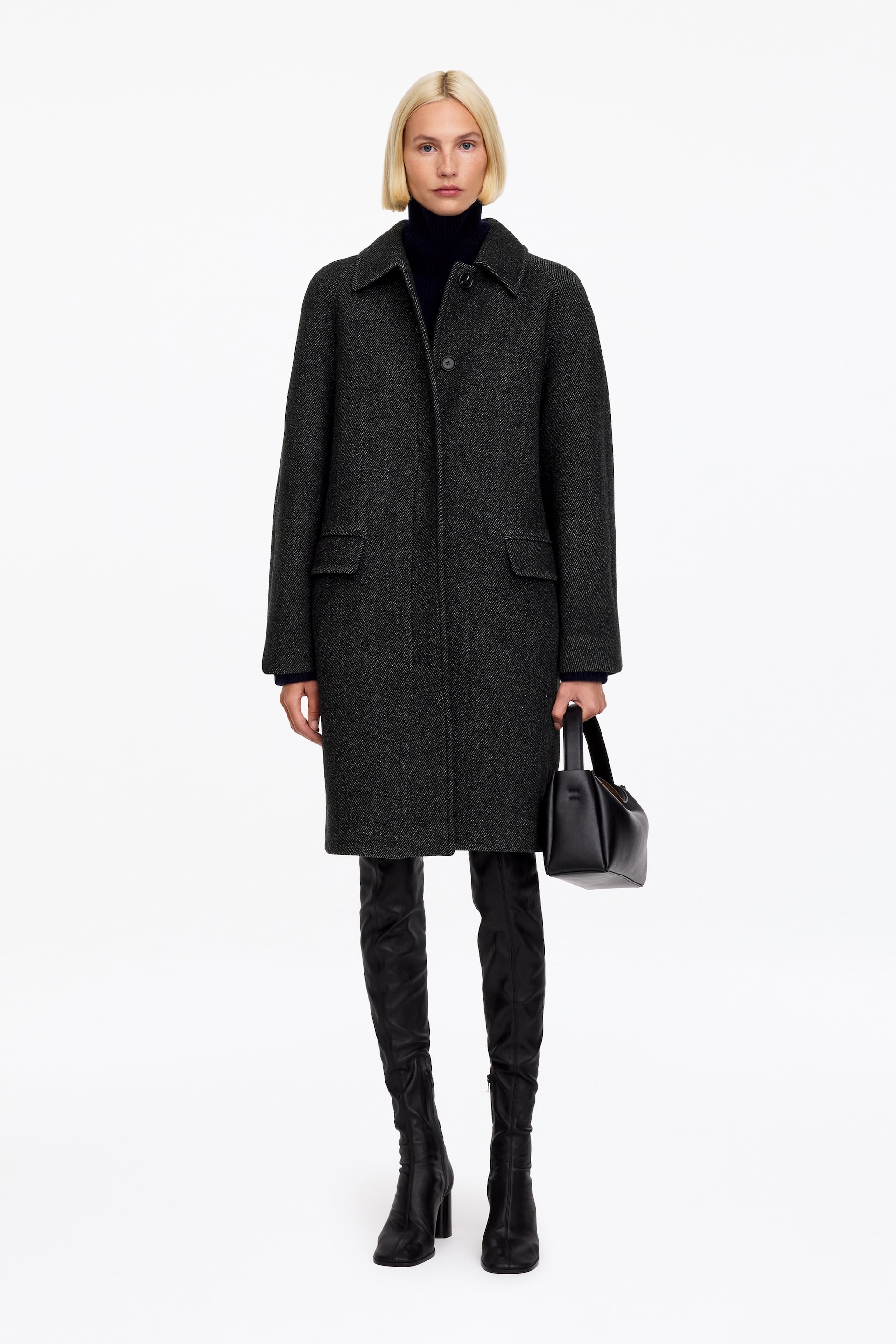 View larger image: The model is wearing a dark grey, textured wool-blend coat featuring a classic collar, front flap pockets, and a knee-length hem. The coat is layered over a dark roll-neck sweater, with black over-the-knee boots and a black leather handbag carried in one hand.