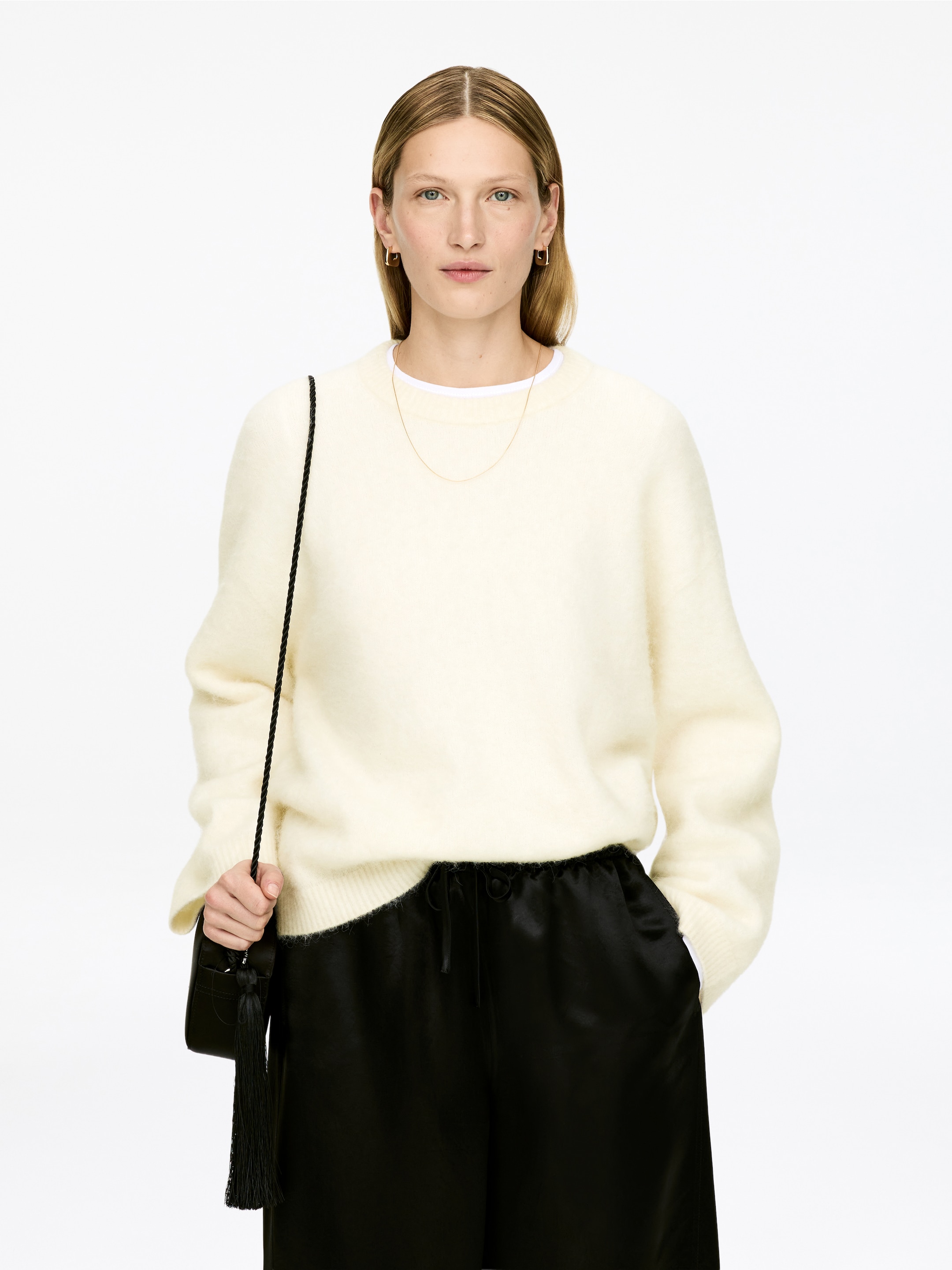 Grösseres Bild ansehen: Ein cremefarbener, gebürsteter Strickpullover mit Rundhalsausschnitt, der ein weißes T-Shirt zeigt, das am Ausschnitt darunter geschichtet ist, wird von vorne präsentiert. Er zeichnet sich durch eine lockere Passform mit gerippten Bündchen und Saum aus und wird mit schwarzen, fließenden Kordelzughosen kombiniert.