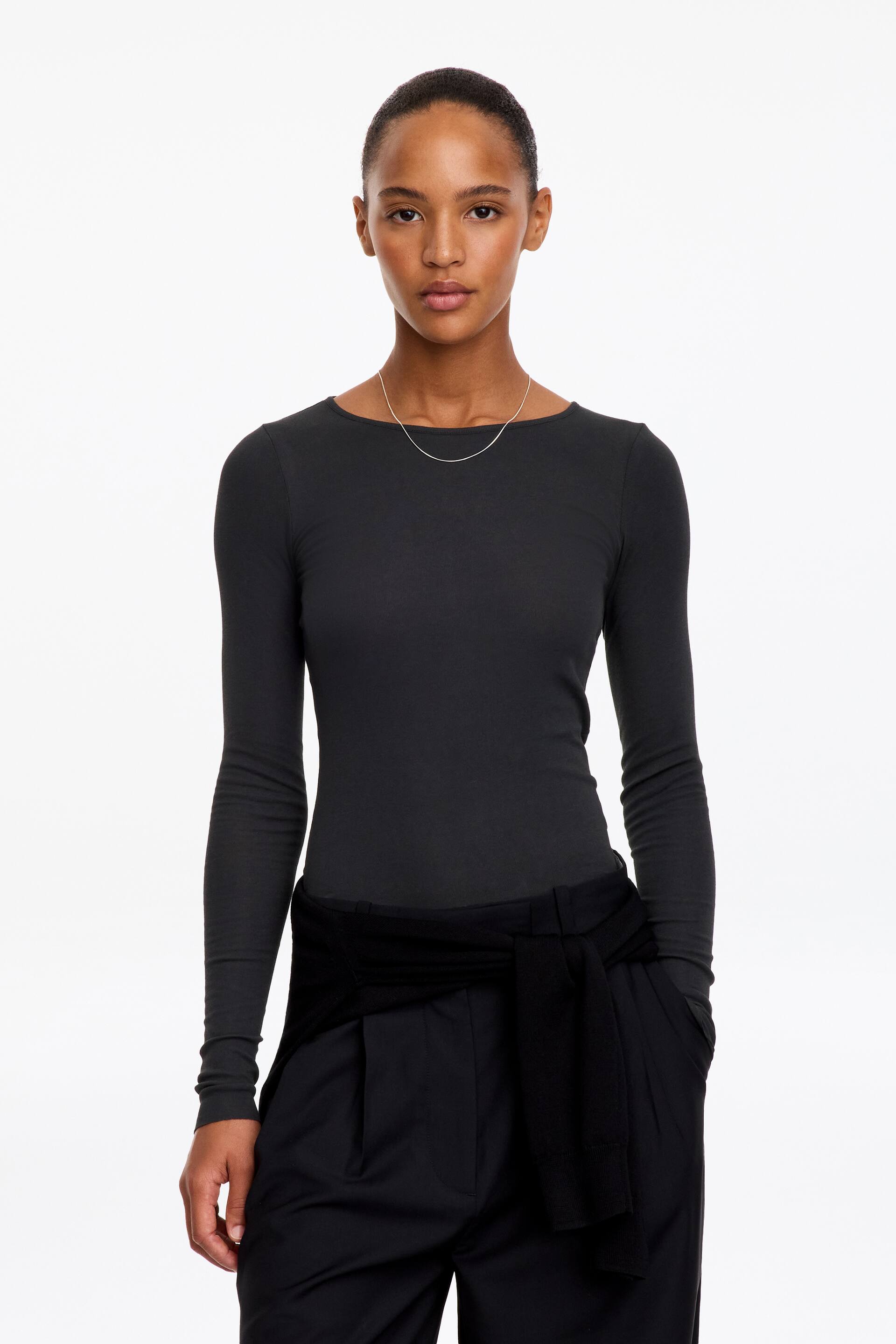 Größeres Bild anzeigen: Das Model trägt ein dunkelgraues, langärmliges Oberteil mit Rundhalsausschnitt und schmaler Passform. Das Oberteil ist in eine schwarze Hose gesteckt, die mit einem schwarzen Strickteil um die Taille gebunden ist.