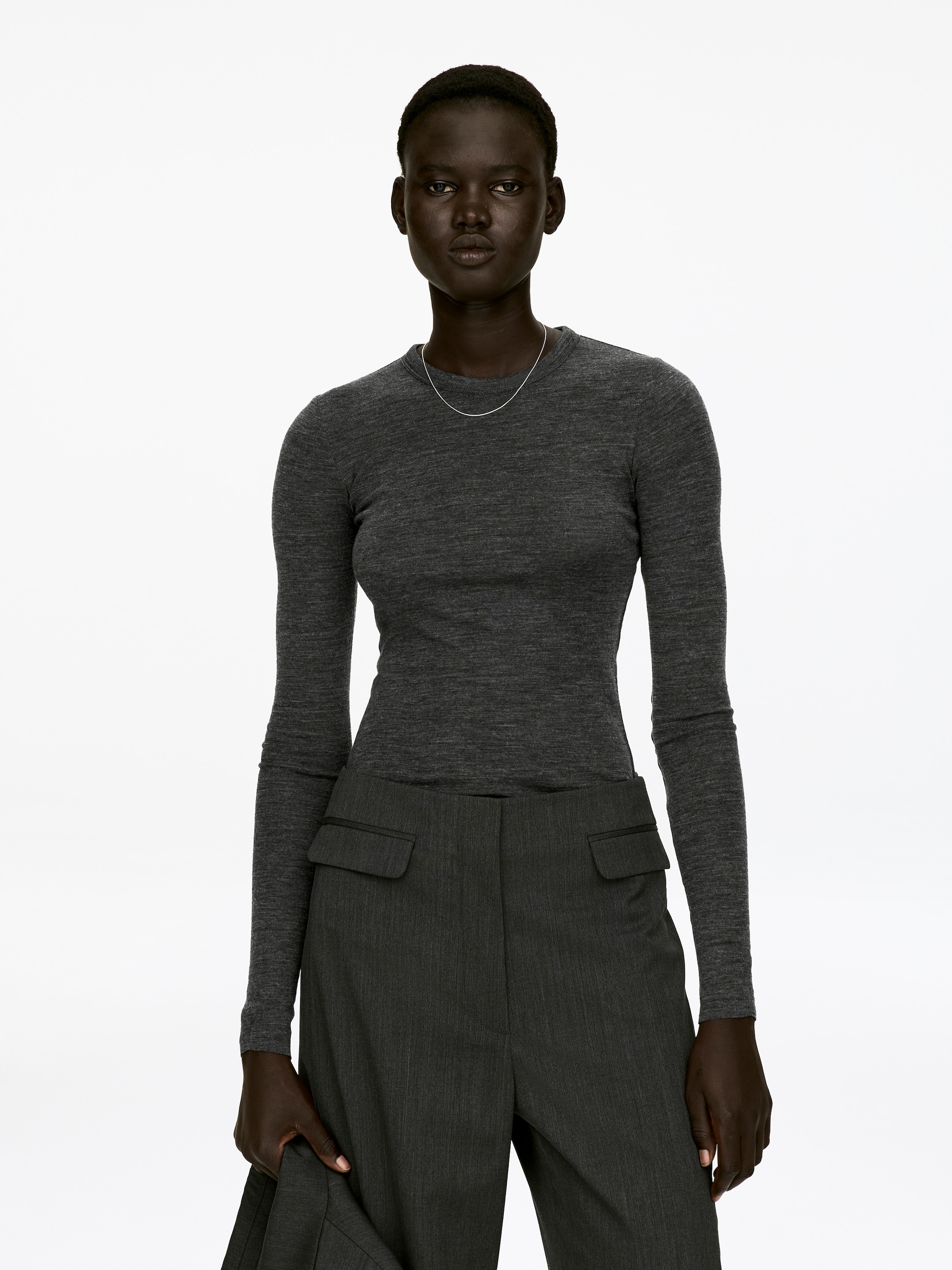 Größeres Bild anzeigen: Ein dunkelgraues, eng anliegendes Langarmshirt mit roh geschnittenen Kanten am Saum und an den Ärmelenden wird von dem Model getragen. Eine dünne silberfarbene Halskette liegt auf dem Ausschnitt des Oberteils, und das Model trägt ebenfalls dunkelgraue Hosen.