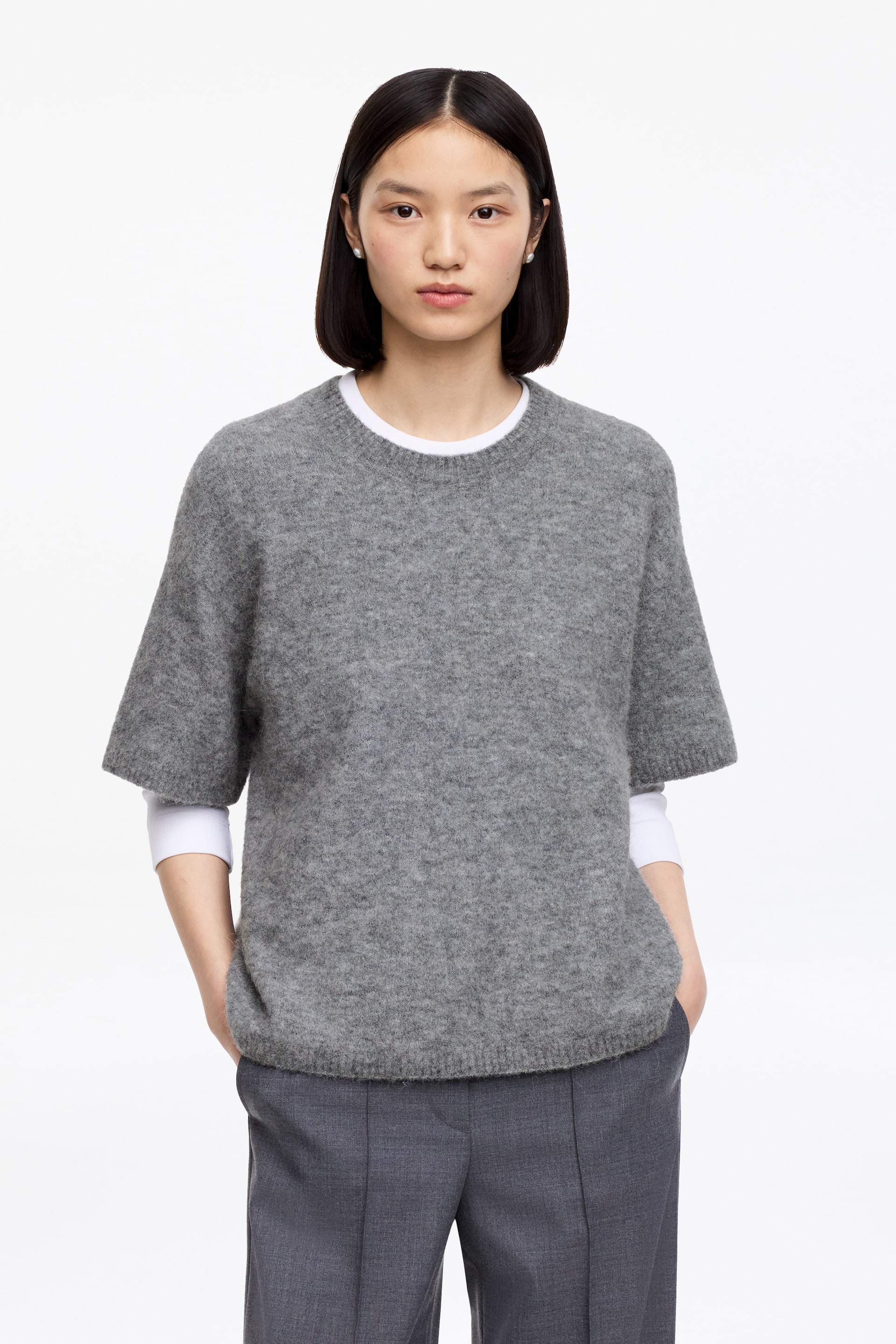 Преглед на по-голямо изображение: Сив пуловер с мъхеста текстура, който се отличава с кръгло деколте, къси ръкави, завършващи над лакътя, и рипсена подгъвка. Носен е на пластове върху бяла блуза с дълги ръкави, като бялата материя е видима на деколтето и се простира от ръкавите.