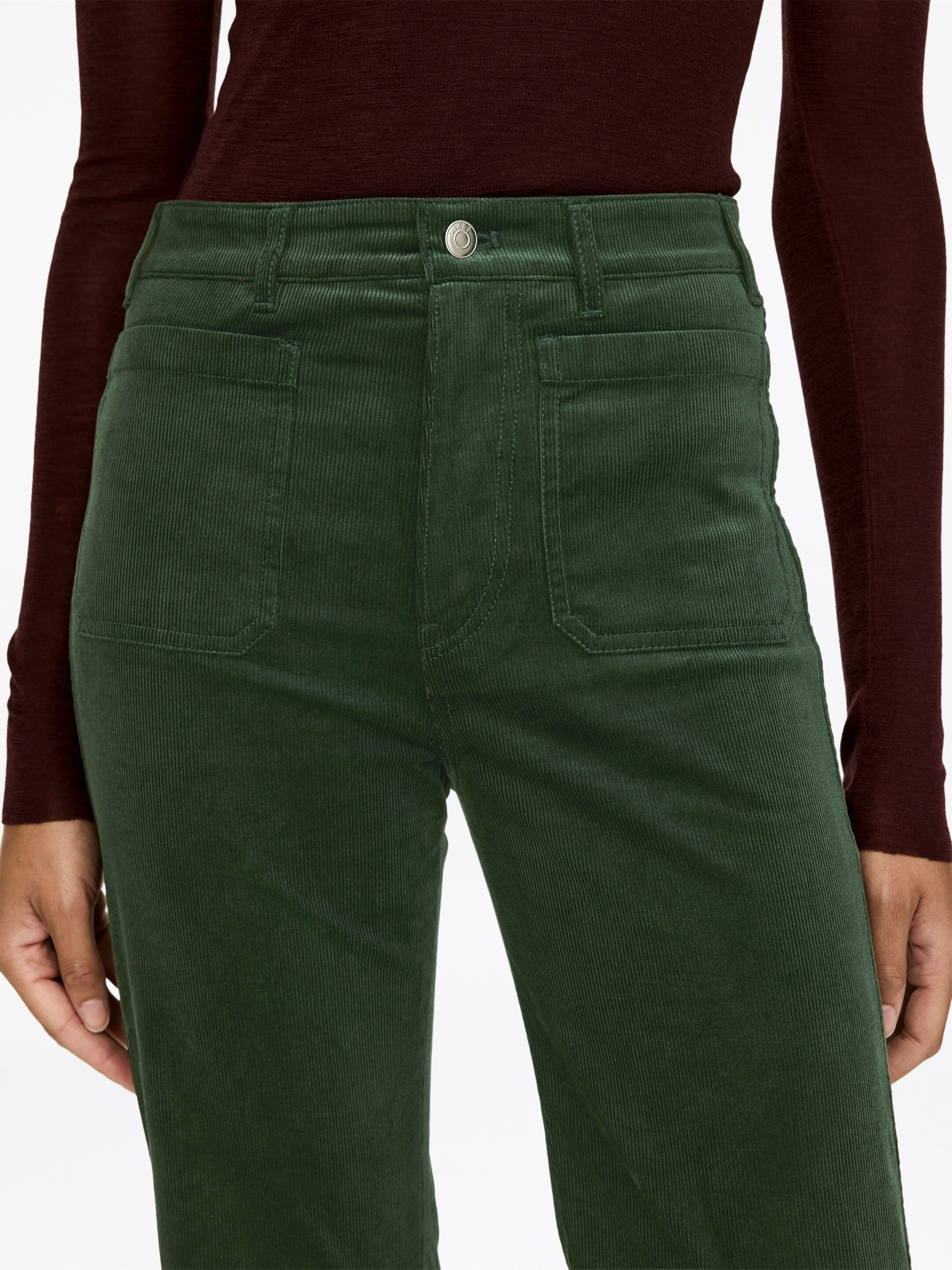 Ver imagen más grande: Pantalones de pana verde oscuro presentan un diseño de talla alta, completos con trabillas para el cinturón, un cierre de botón plateado y una bragueta con cremallera. Dos bolsillos de parche rectangulares son visibles en la parte delantera, y los pantalones muestran su característica textura acanalada.