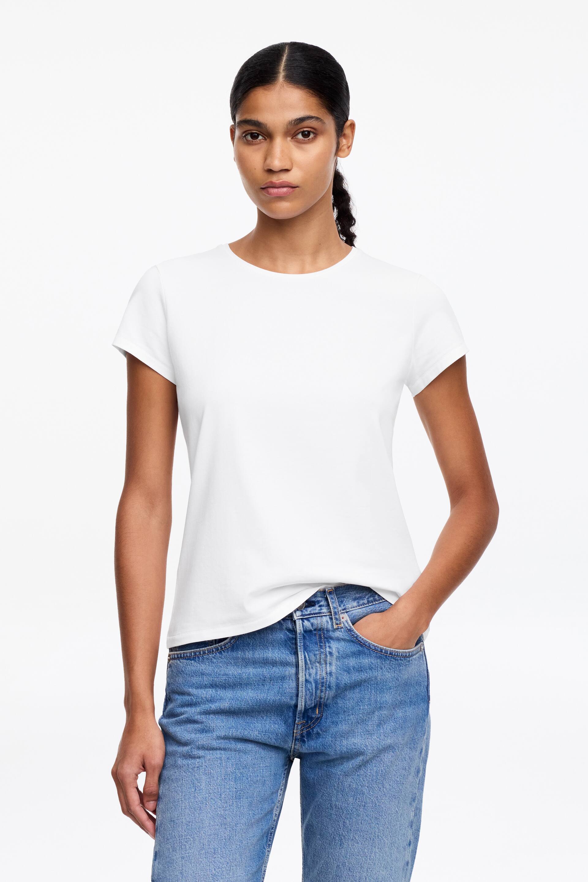 Visa större bild: En vit kortärmad t-shirt med rund halsringning och en något figurnära silhuett. Den bärs med fållen instoppad i blå jeans på ena sidan och hängande på den andra.