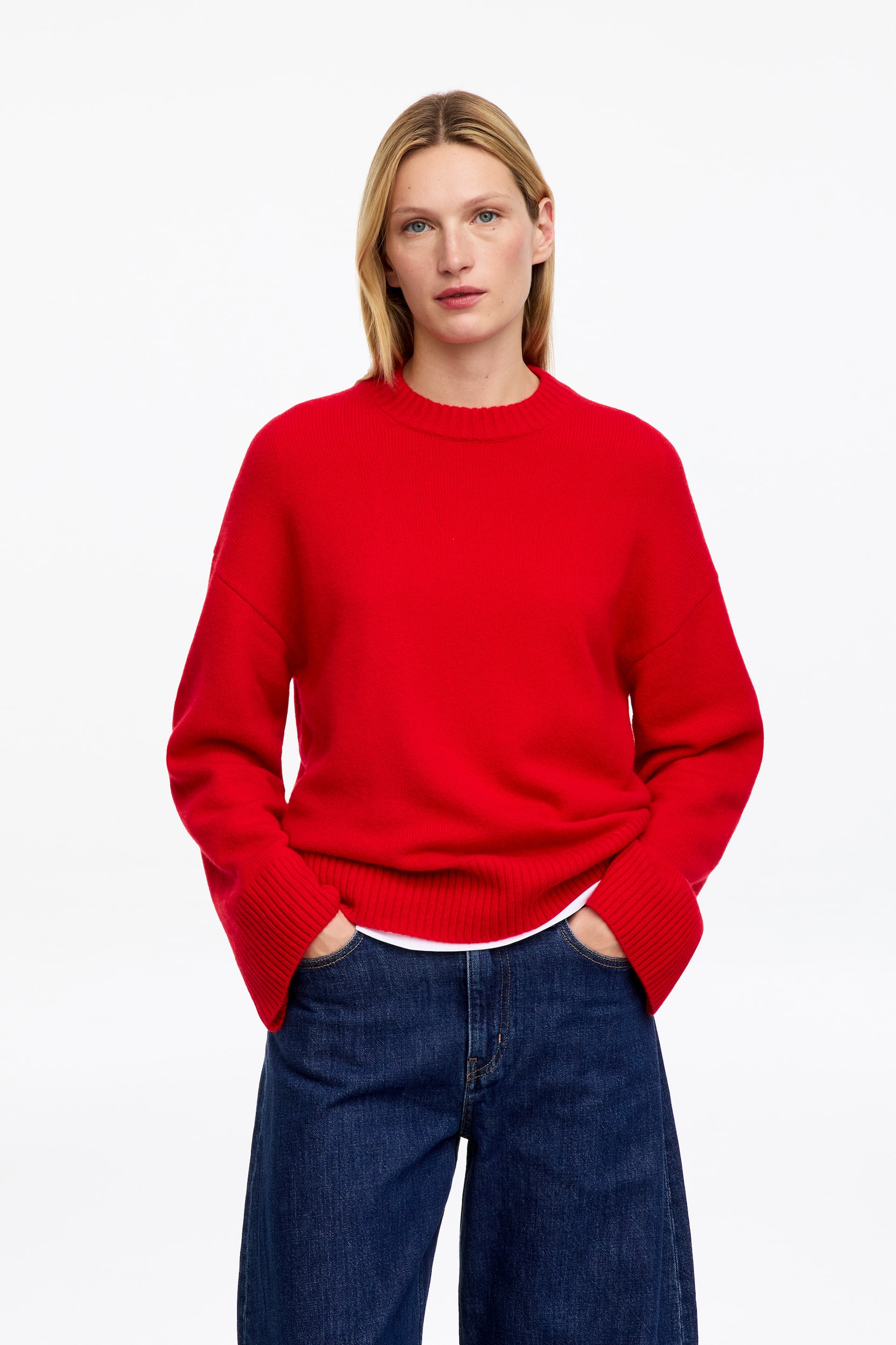 Visualizza immagine più grande: Un maglione rosso lavorato a maglia con girocollo, spalle scese, ampi polsini a coste e orlo a coste è indossato dalla modella. Il maglione è abbinato a una T-shirt bianca ed è indossato con jeans blu scuro, mentre la modella tiene le mani nelle tasche anteriori.