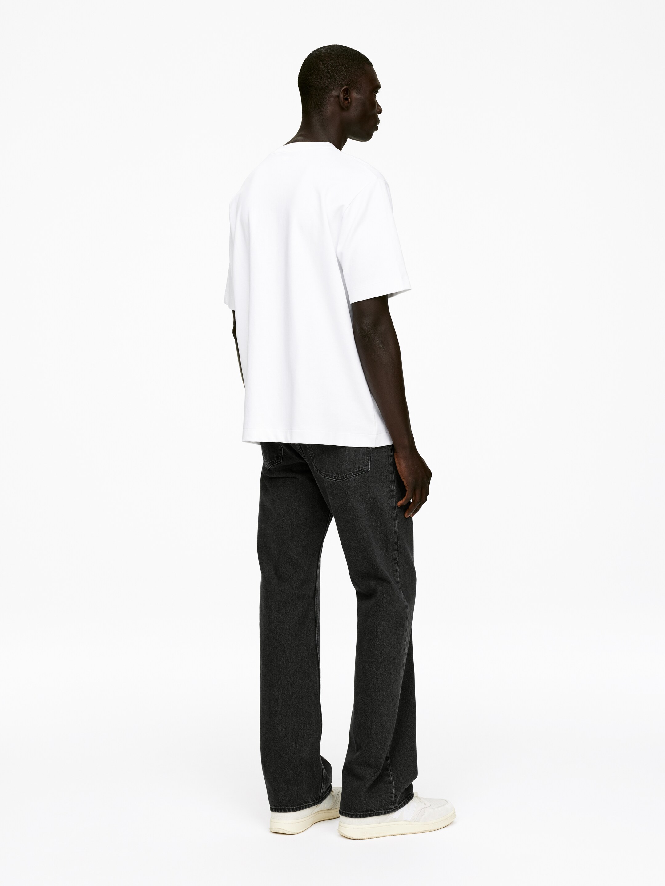 Ingrandisci l'immagine: Una t-shirt bianca con scollo a girocollo, caratterizzata da maniche corte e una vestibilità rilassata, è indossata. È abbinata a jeans grigio scuro, a gamba dritta e vestibilità rilassata, e sneakers bianche a taglio basso. I prodotti sono presentati da un'angolazione posteriore, evidenziando il drappeggio casual della t-shirt e il taglio ampio dei jeans.