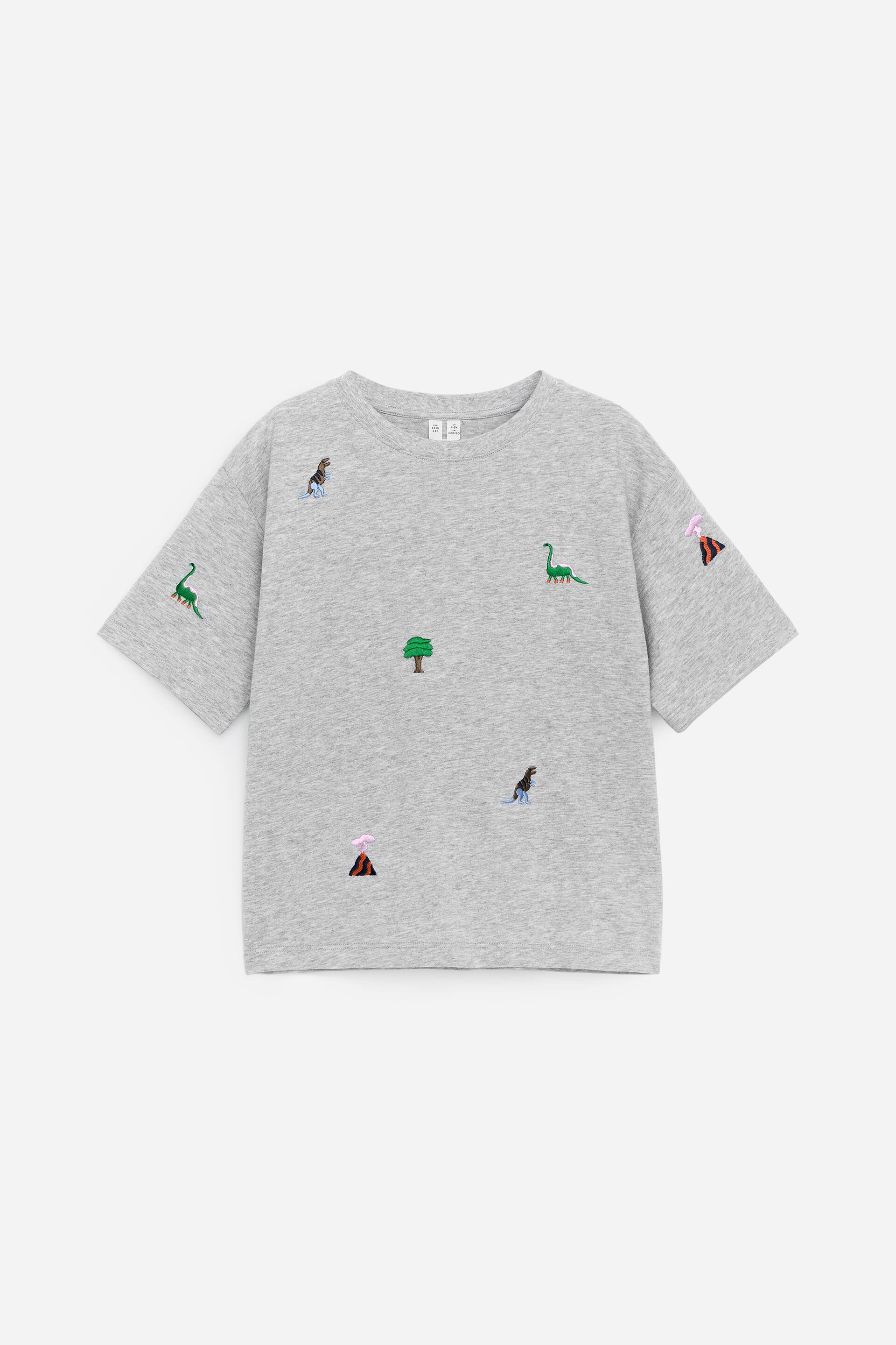 Visualizza immagine più grande: Una maglietta grigia a maniche corte con scollo a girocollo presenta il suo fronte, caratterizzata da vari motivi ricamati sparsi sulla sua superficie. Dinosauri dal collo lungo verdi, dinosauri marrone scuro, alberi verdi e vulcani con fumo rosa decorano la maglietta.