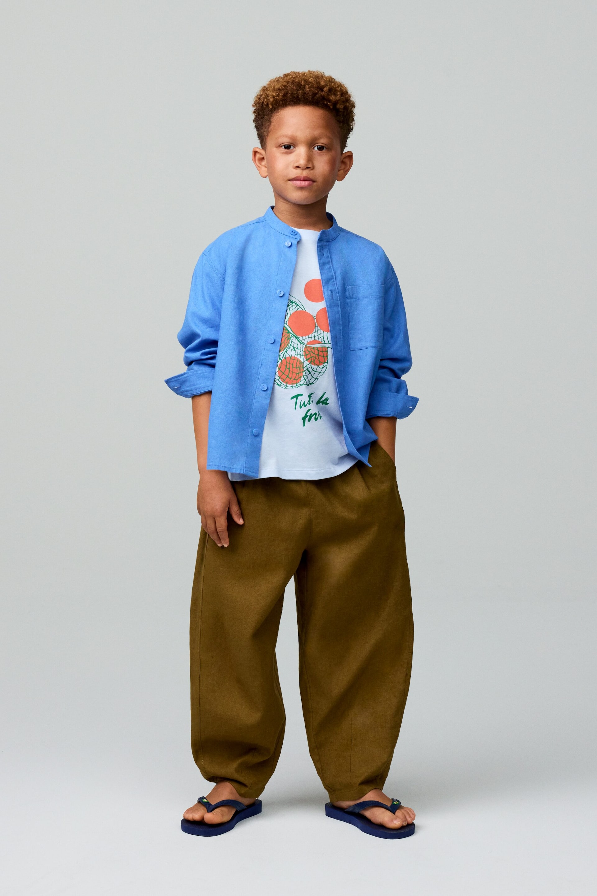 Zobraziť väčší obrázok: Modrá košeľa s dlhými rukávmi a stojatým golierom, so zrolovanými rukávmi a náprsným vreckom, je nosená otvorená cez biele tričko s oranžovou a sieťovou grafikou. Tento outfit dopĺňajú voľné olivovo zelené nohavice a námornícke modré žabky.