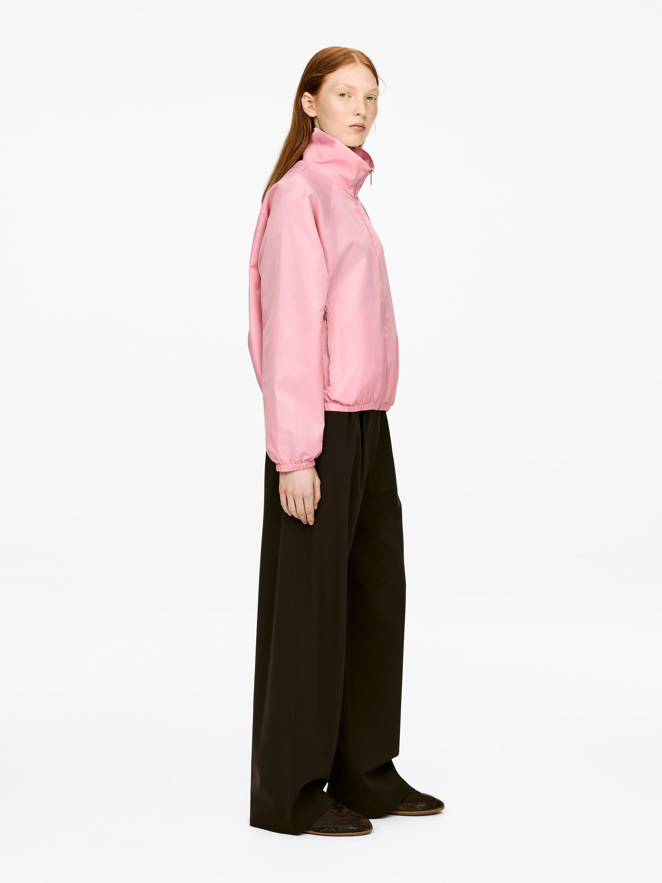 Vis større bilde: En sporty rosa jakke i nylon med høy krage, metalldetaljer på glidelåsen, og elastiske mansjetter og nederkant bæres av modellen. Modellen står og har på seg jakken over mørke bukser.