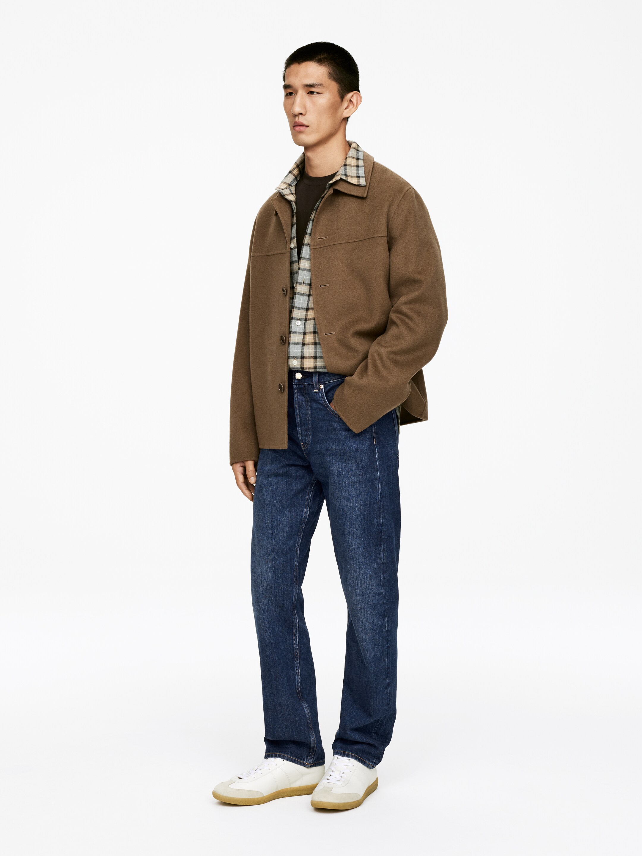 Visa större bild: En fullständig outfit presenteras, med en brun jacka med krage och horisontella sömmar över bröstet, bärs öppen över en ljus rutig skjorta och en mörk t-shirt med rund hals. Ensemblen kompletteras med mörka raka jeans och vita snörskor med ljusgrå mockadetaljer och gummisulor. Kläderna är vinklade något åt höger, vilket visar fram- och sidoprofilerna.