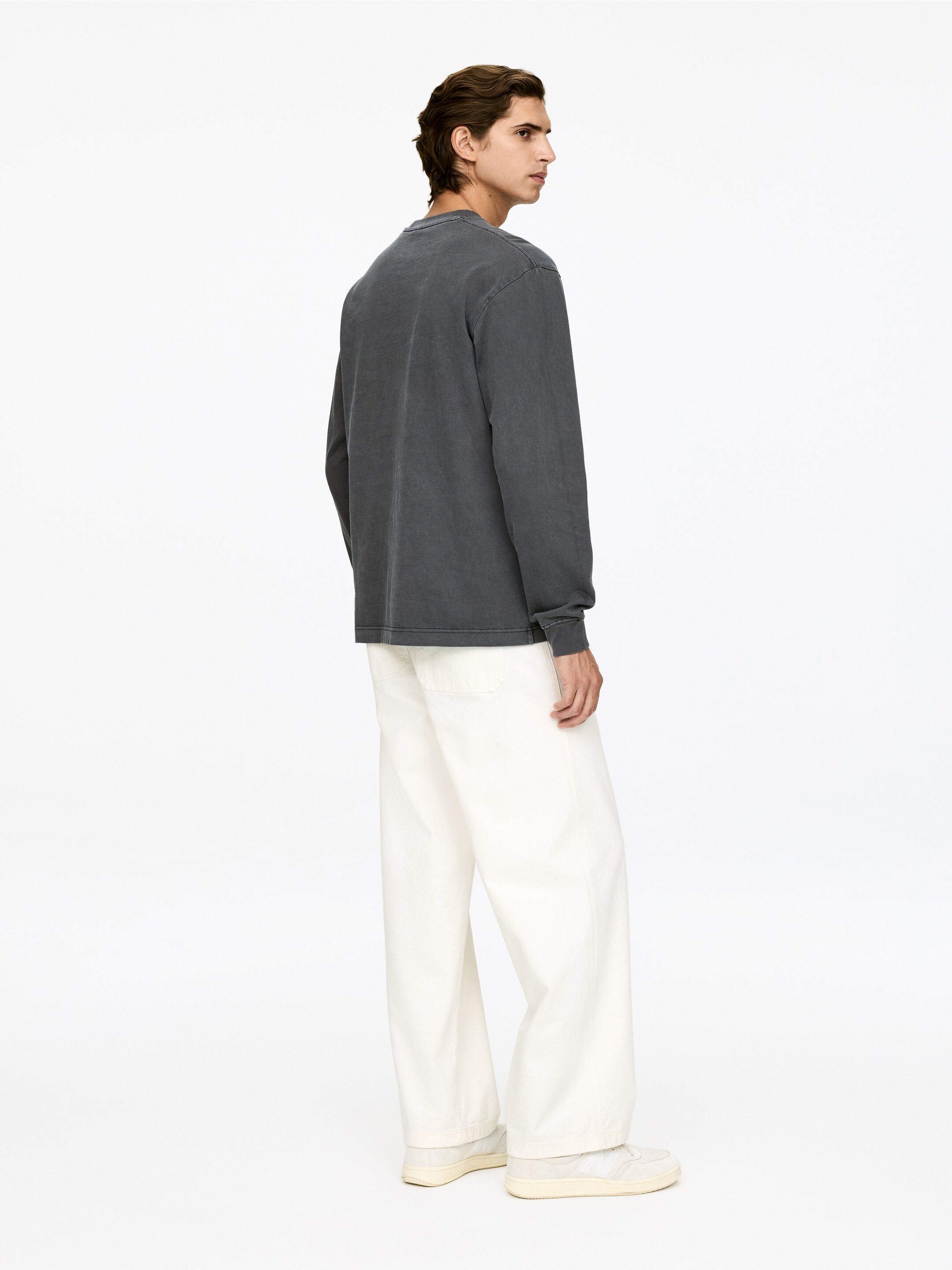 View larger image: A dark gray long-sleeved crew-neck shirt is being presented from the back and side, featuring a relaxed fit and a faded wash. It is being styled with off-white wide-leg trousers that are pooling over cream sneakers.