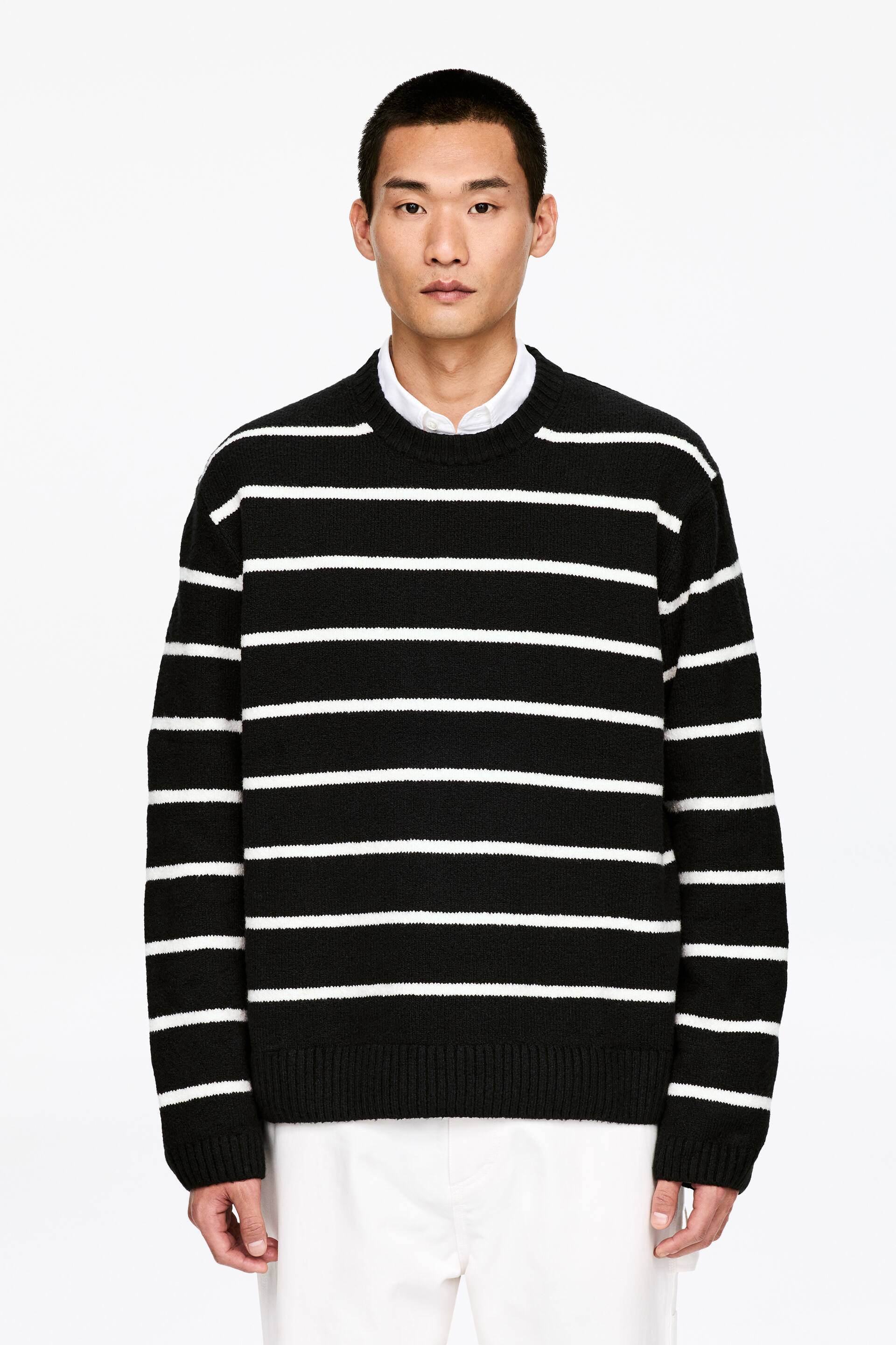 Größeres Bild ansehen: Ein schwarz-weiß gestreifter Pullover ist nach vorne gerichtet und zeigt horizontale weiße Streifen auf einem schwarzen Strickstoff. Er ist mit einem Rundhalsausschnitt, gerippten Bündchen und einem gerippten Saum gestaltet und wird über einem weißen Hemd mit Kragen getragen.