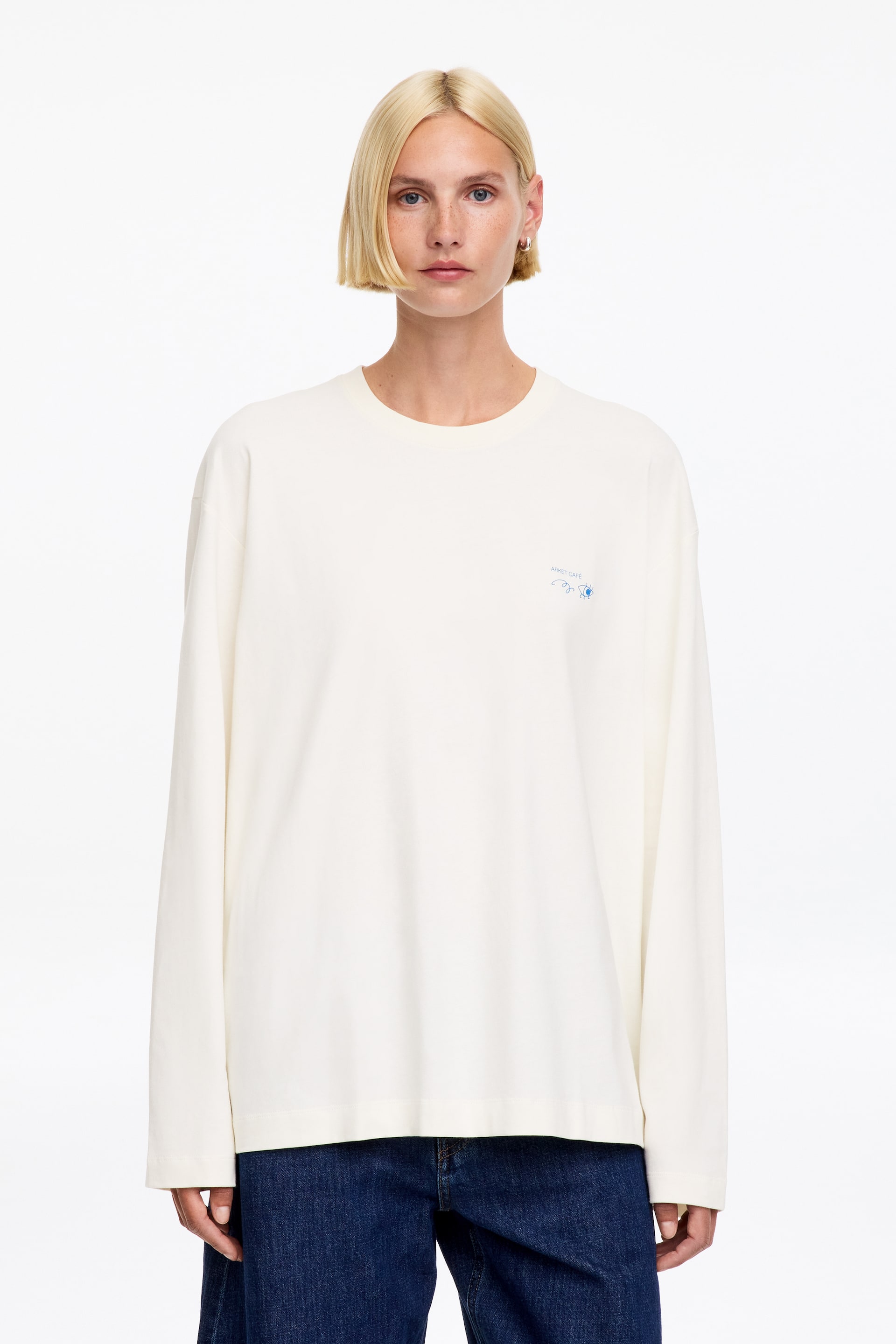 Größeres Bild ansehen: Das cremefarbene, oversized Langarmshirt verfügt über einen Rundhalsausschnitt, eine kleine blaue Stickerei auf der linken Brust und einen geraden Saum, der eine entspannte Passform zeigt.