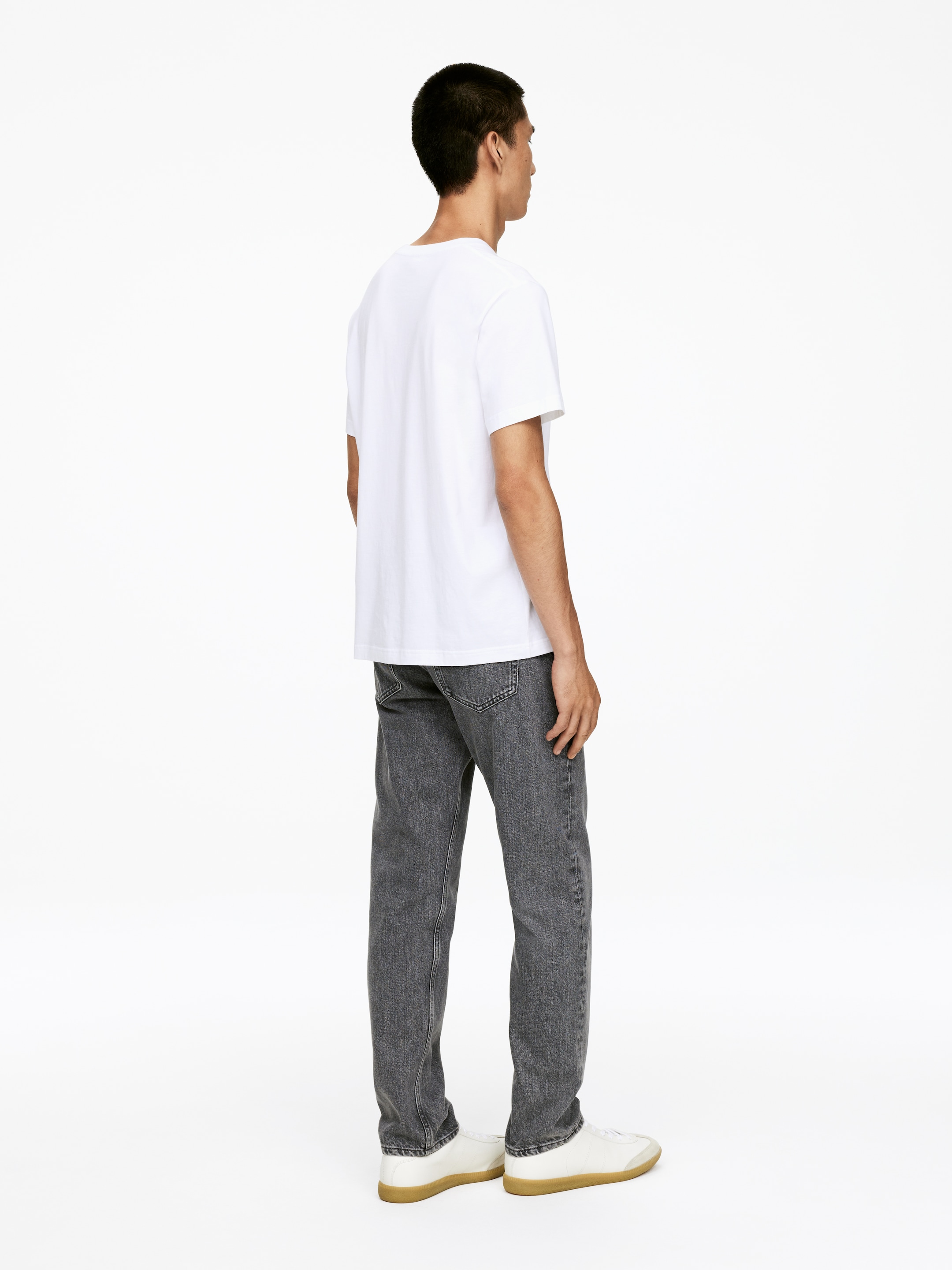 Grösseres Bild ansehen: Ein weißes T-Shirt mit Rundhalsausschnitt wird getragen, das einen entspannten Schnitt und kurze Ärmel aufweist, kombiniert mit grauen, gewaschenen Jeans mit geradem Bein, die ebenfalls eine entspannte Silhouette präsentieren. Weiße Low-Top-Sneaker mit hellbraunen Gummisohlen vervollständigen das Outfit, alles aus einem dreiviertel Rückwinkel betrachtet.