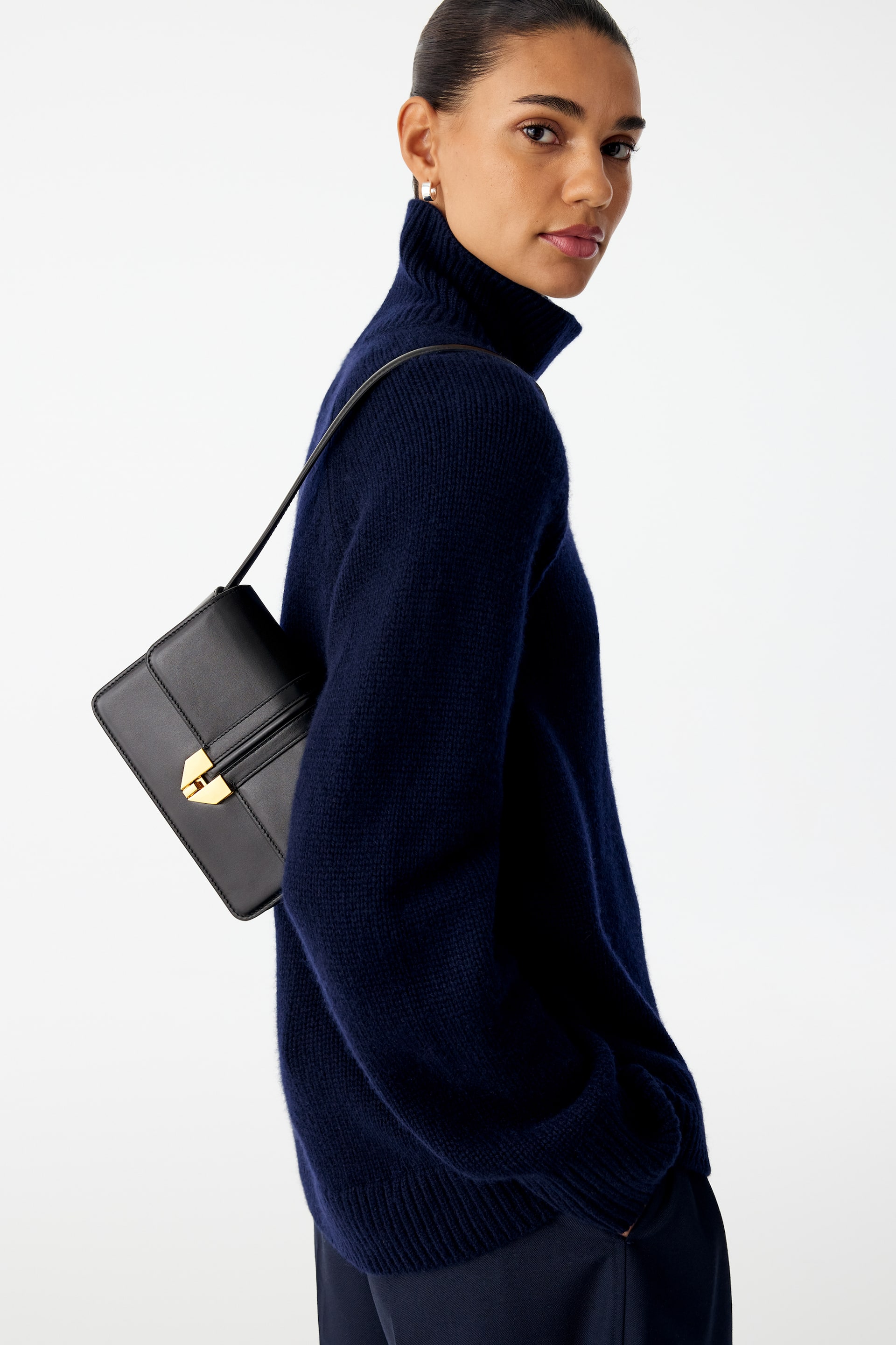 Größeres Bild ansehen: Eine schwarze Leder-Umhängetasche mit kantiger Silhouette und goldfarbenem Verschluss wird über der Schulter des Models getragen. Der schmale Riemen der Tasche liegt auf einem dunkelblauen Rollkragenpullover.