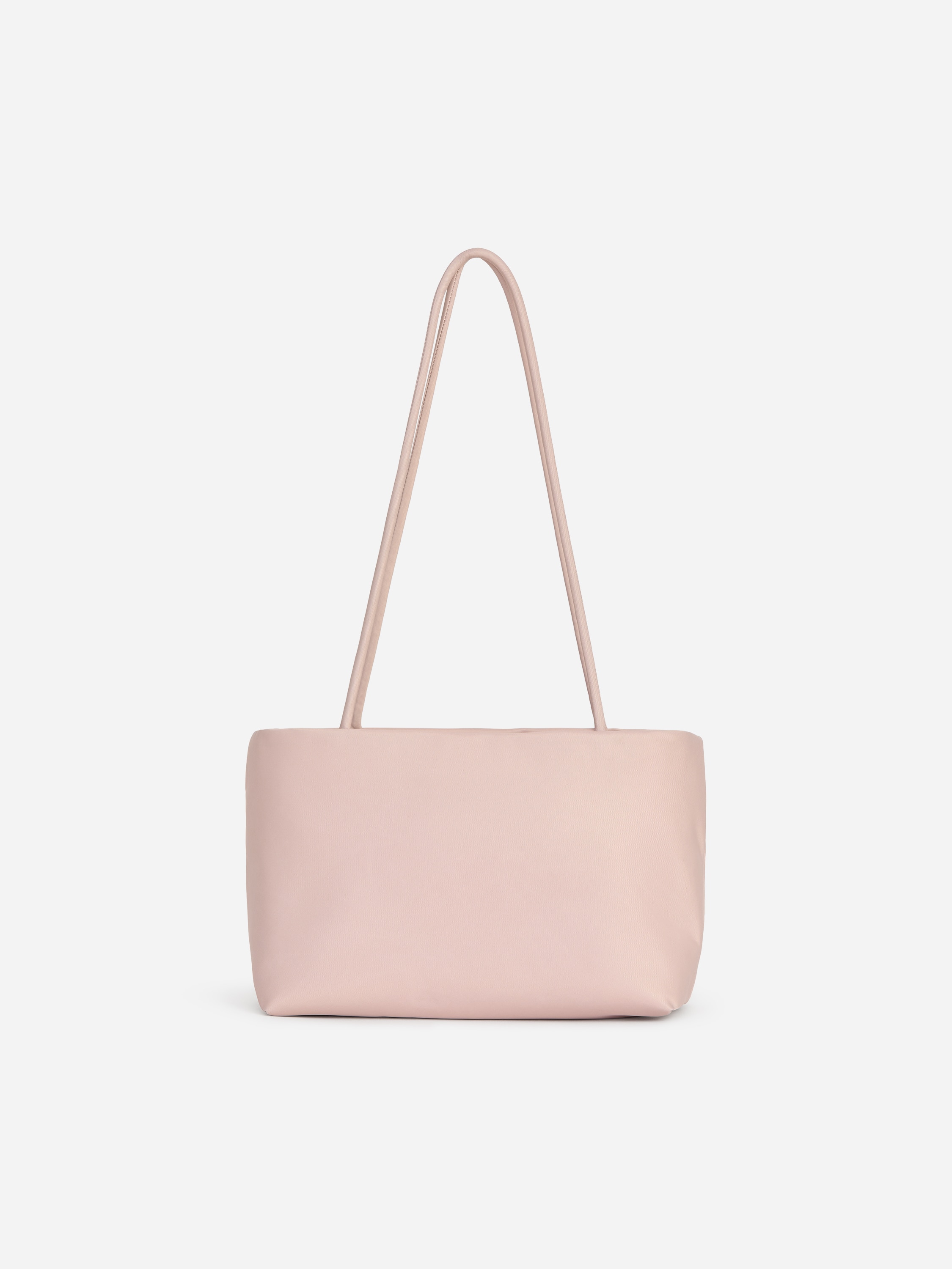 Een lichtroze, langwerpige boxy nylon totebag met slanke handgrepen wordt gedragen door het model, dat hem aan de hengsels vasthoudt. De tas wordt gecombineerd met een zwarte leren jas met hoge kraag en een zwarte broek.
