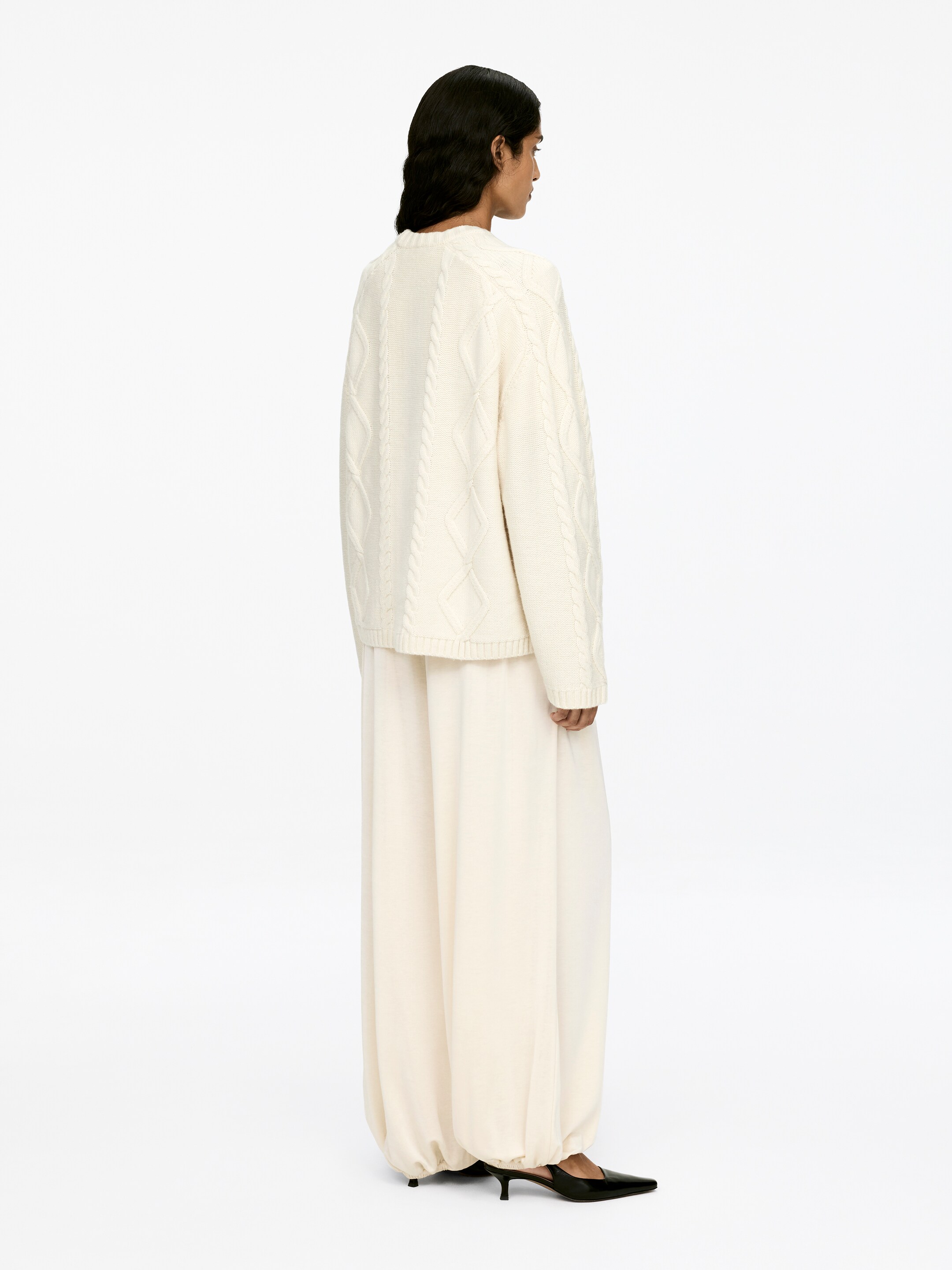 View larger image: A cream cable knit jumper, featuring a crew-neck, long sleeves, and ribbed trims, is being worn by the model. The relaxed-fit jumper is styled with wide, coordinating trousers.