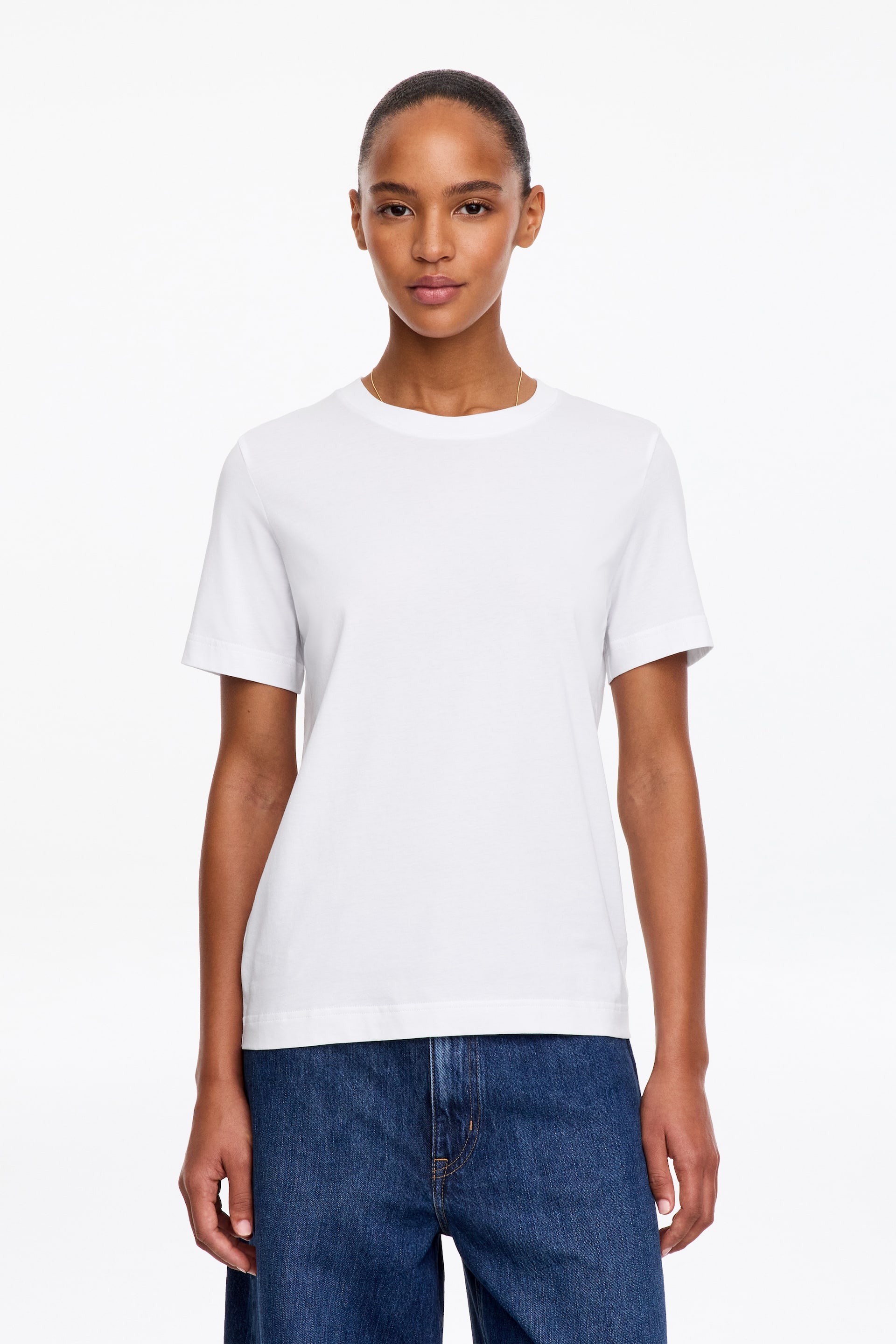 Ver imagem maior: A modelo veste uma t-shirt branca de gola redonda, com mangas curtas e bainha direita. A t-shirt é combinada com jeans de ganga azul.