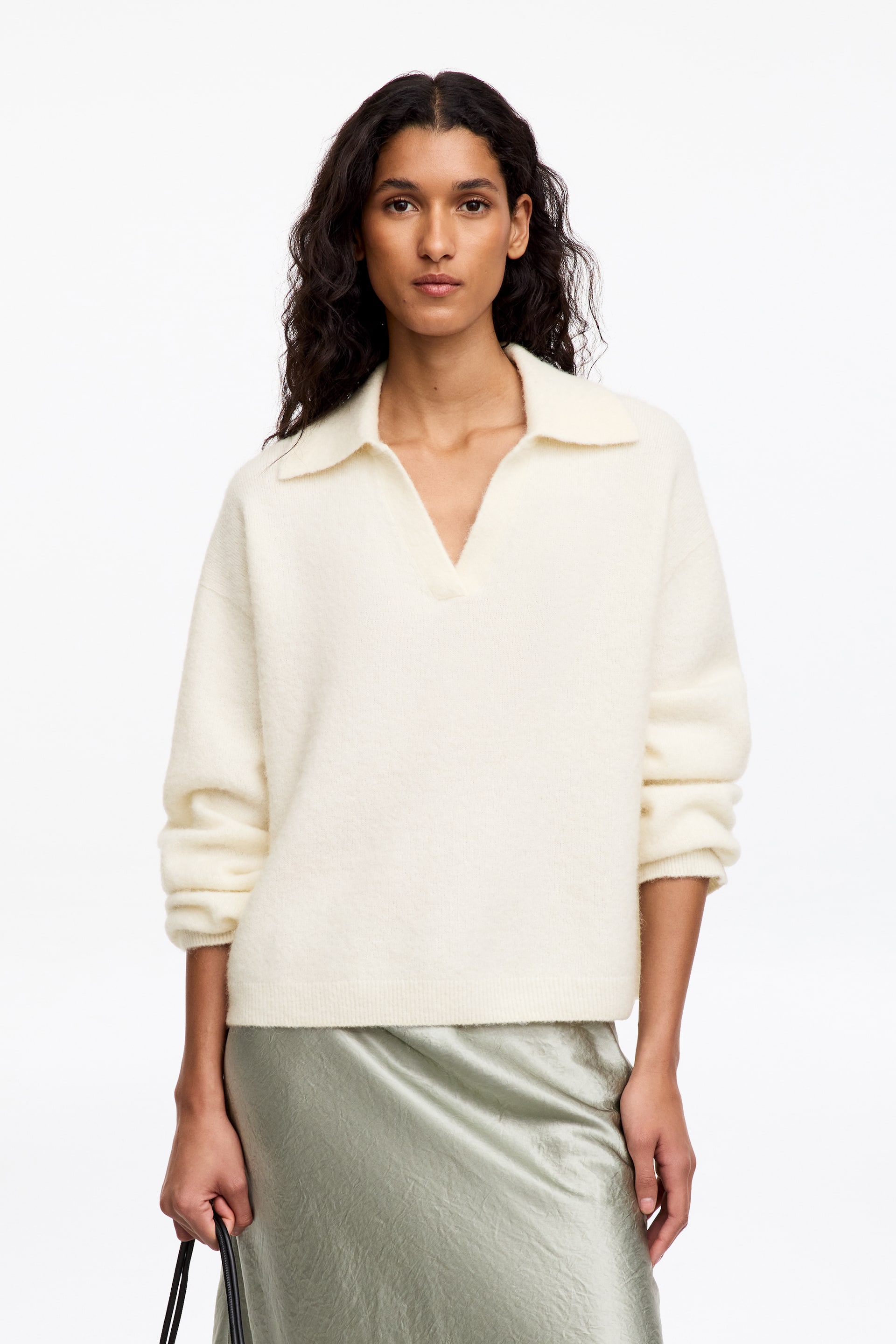 Agrandir l'image: Un pull en maille crème à la texture duveteuse est porté par le mannequin. Il présente un col polo côtelé avec une encolure en V, une coupe ample et des manches retroussées, associé à une jupe vert clair.