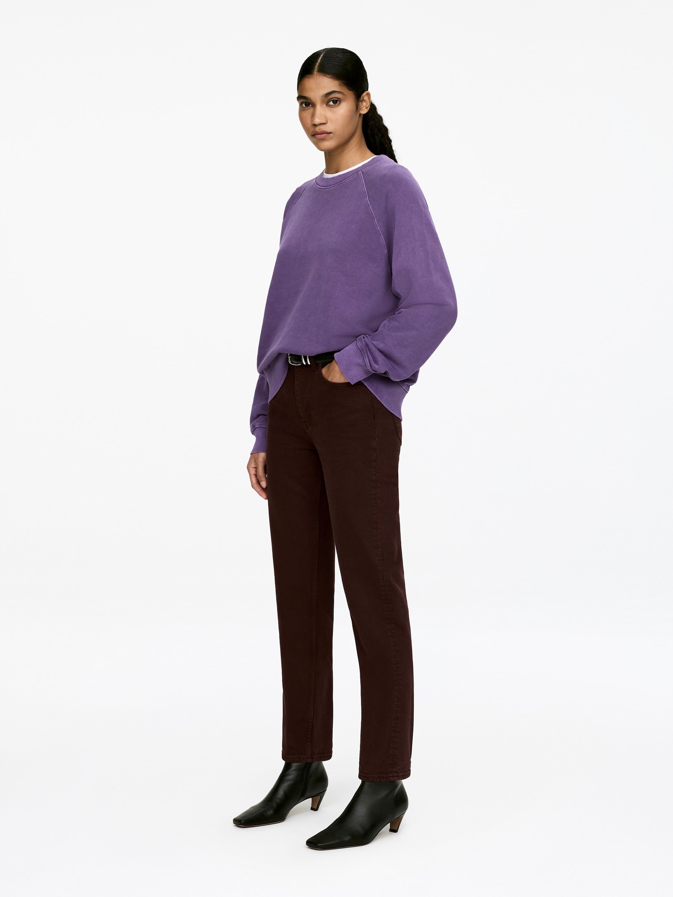 Grösseres Bild ansehen: Ein lila Sweatshirt mit Rundhalsausschnitt, das über Raglanärmel und gerippte Abschlüsse verfügt, wird vom Model über einem weissen T-Shirt getragen, wobei eine Hand des Models in einer Hosentasche ruht.