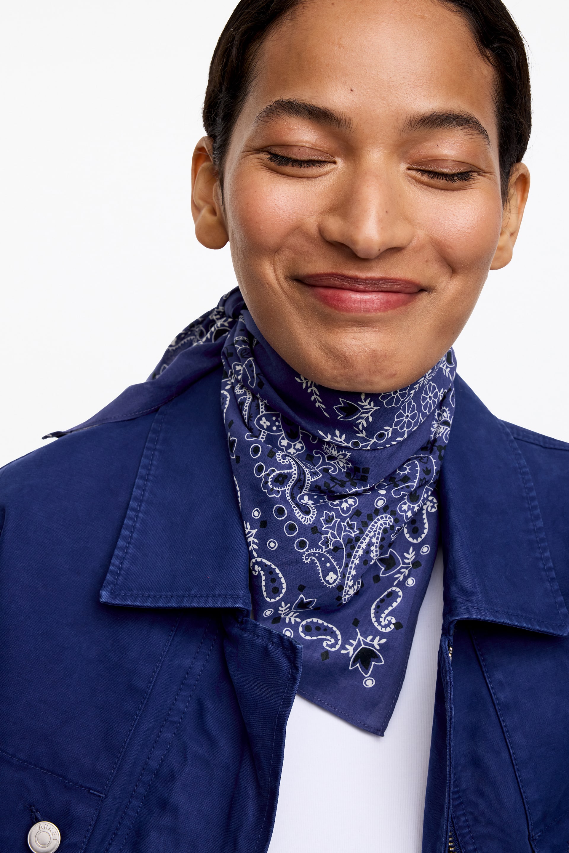 View larger image: Cotton scarf, dark blue with a white paisley pattern, is worn around the model's neck. It is styled beneath an open blue jacket and displayed over a white top.