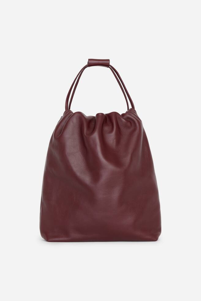 Una borsa in pelle bordeaux intenso Ã¨ posizionata in verticale, mostrando frontalmente una silhouette morbida ispirata a una dustbag. Sottili tracolle a coulisse si estendono verso lâalto dallâapertura superiore arricciata e sono fissate insieme da un laccetto coordinato in pelle. - 1