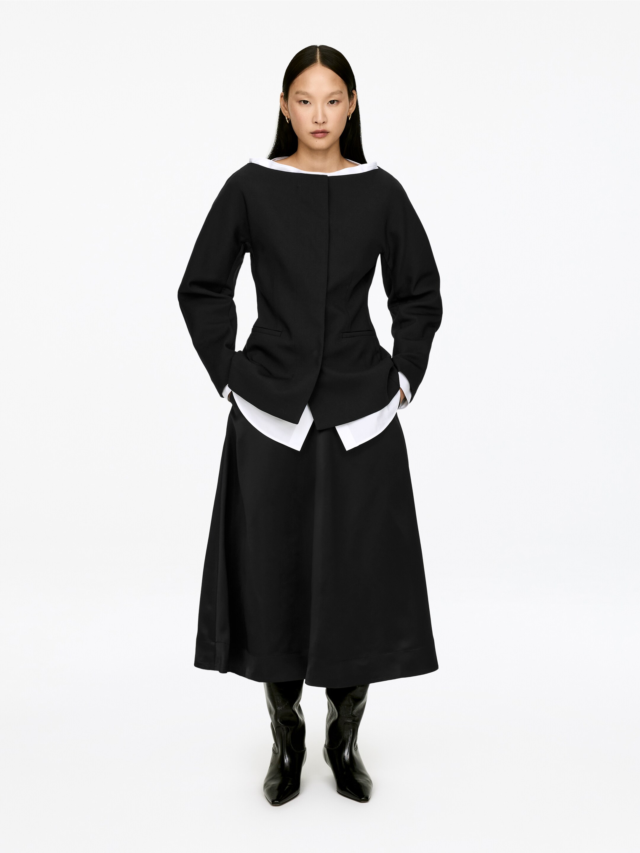 Grösseres Bild ansehen: Ein Ensemble bestehend aus einer schwarzen Jacke und einem Midirock wird aus einem frontalen Winkel präsentiert. Die schwarze Jacke zeigt einen breiten, abgerundeten Ausschnitt, lange Ärmel und einen asymmetrischen Saum mit zwei horizontalen Schlitztaschen, während der abgerundete Kragen und Saum eines weißen Hemdes sichtbar darunter hervorschauen. Diese Jacke bedeckt teilweise einen schwarzen A-Linien-Midirock, der mit schwarzen spitz zulaufenden Stiefeln getragen wird.
