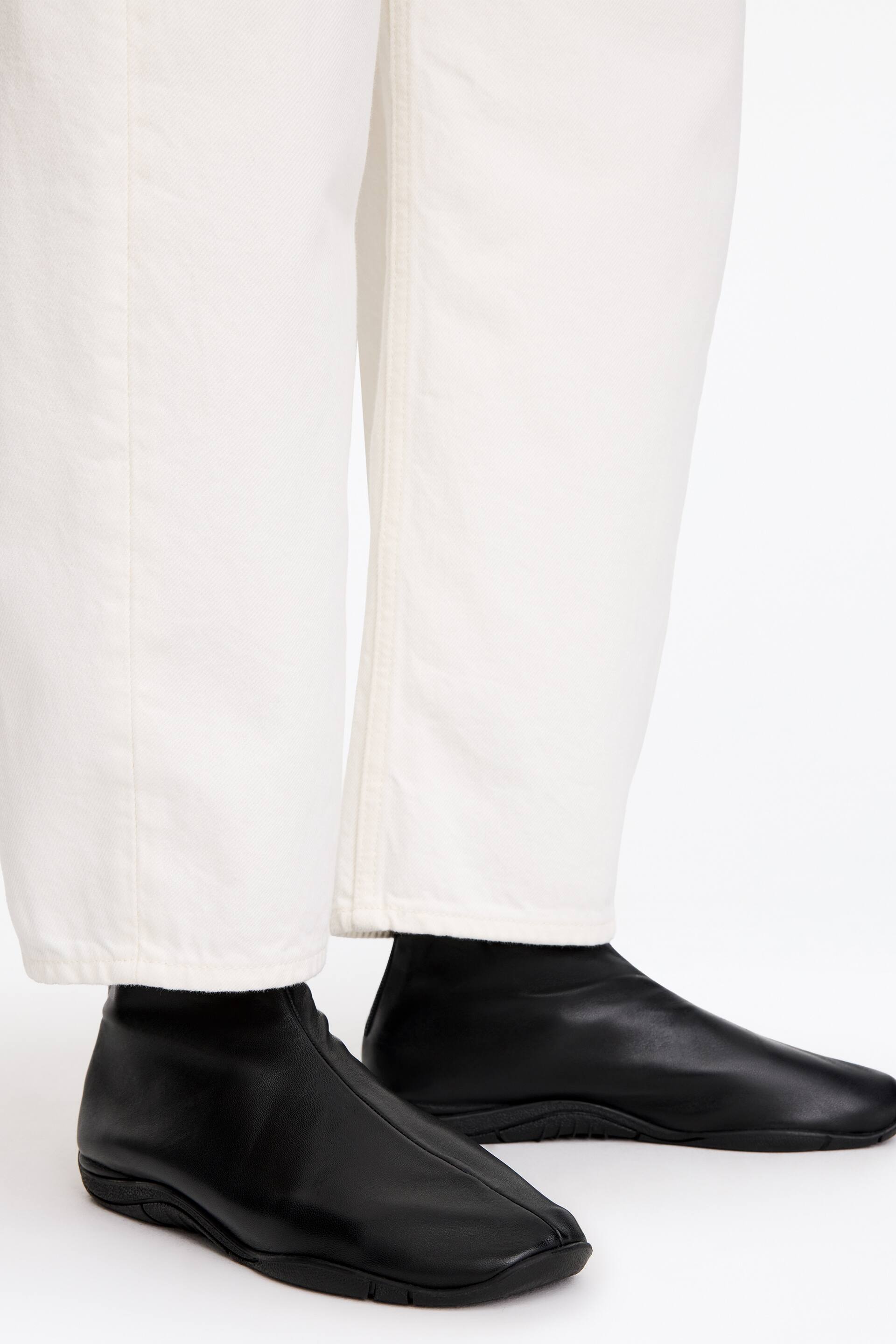 Ver imagen más grande: La modelo lleva botas tobilleras de estilo calcetín en piel negra bajo unos pantalones blancos de pierna ancha. Las botas presentan un diseño ajustado, una costura central que recorre la parte delantera y suelas de goma planas de estilo deportivo.